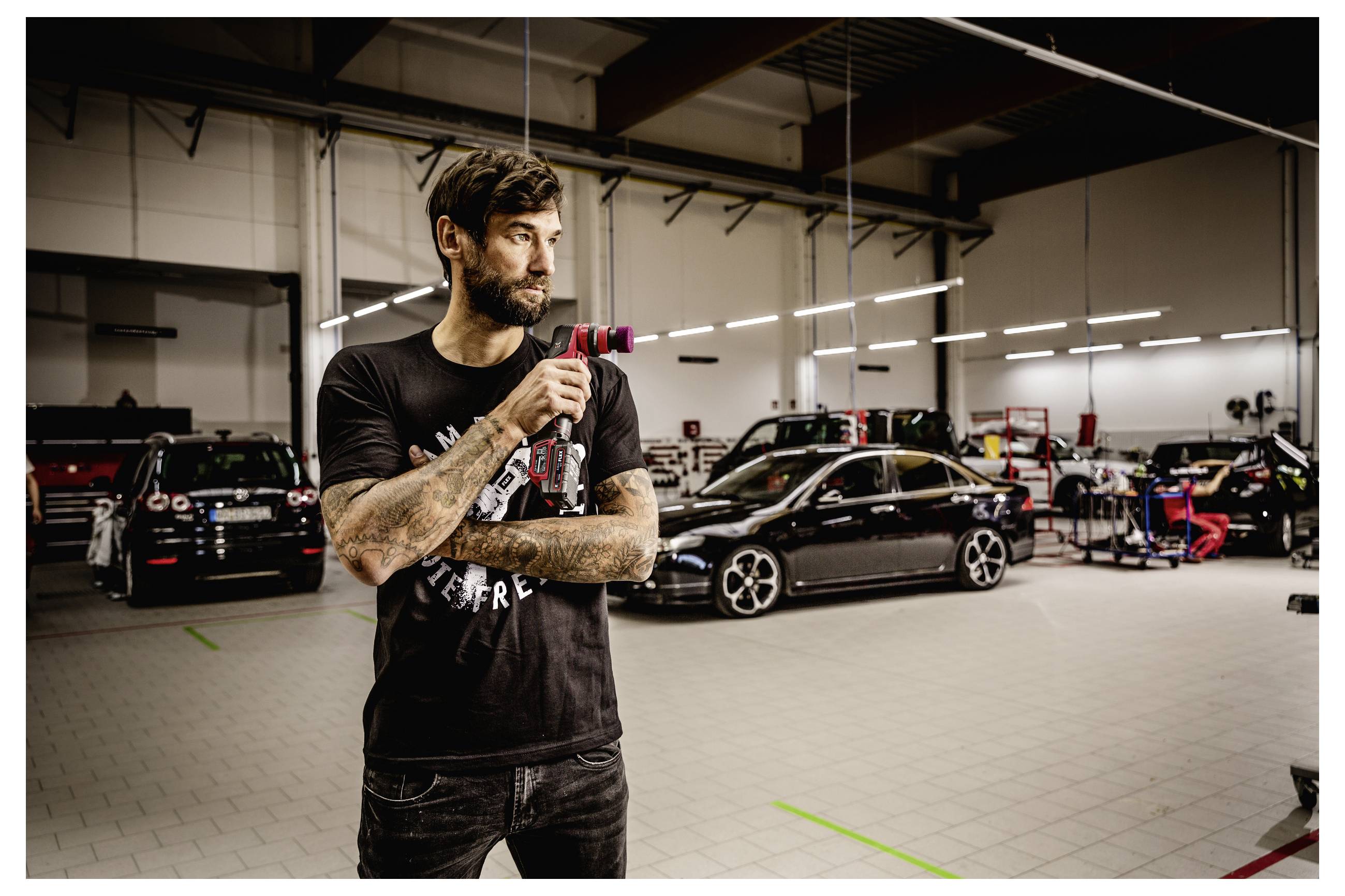 Tätowierter Mann in schwarzem T-Shirt steht in einer Autowerkstatt, hält ein Elektrowerkzeug in der Hand, mit modifizierten Autos und Ausrüstung im Hintergrund.