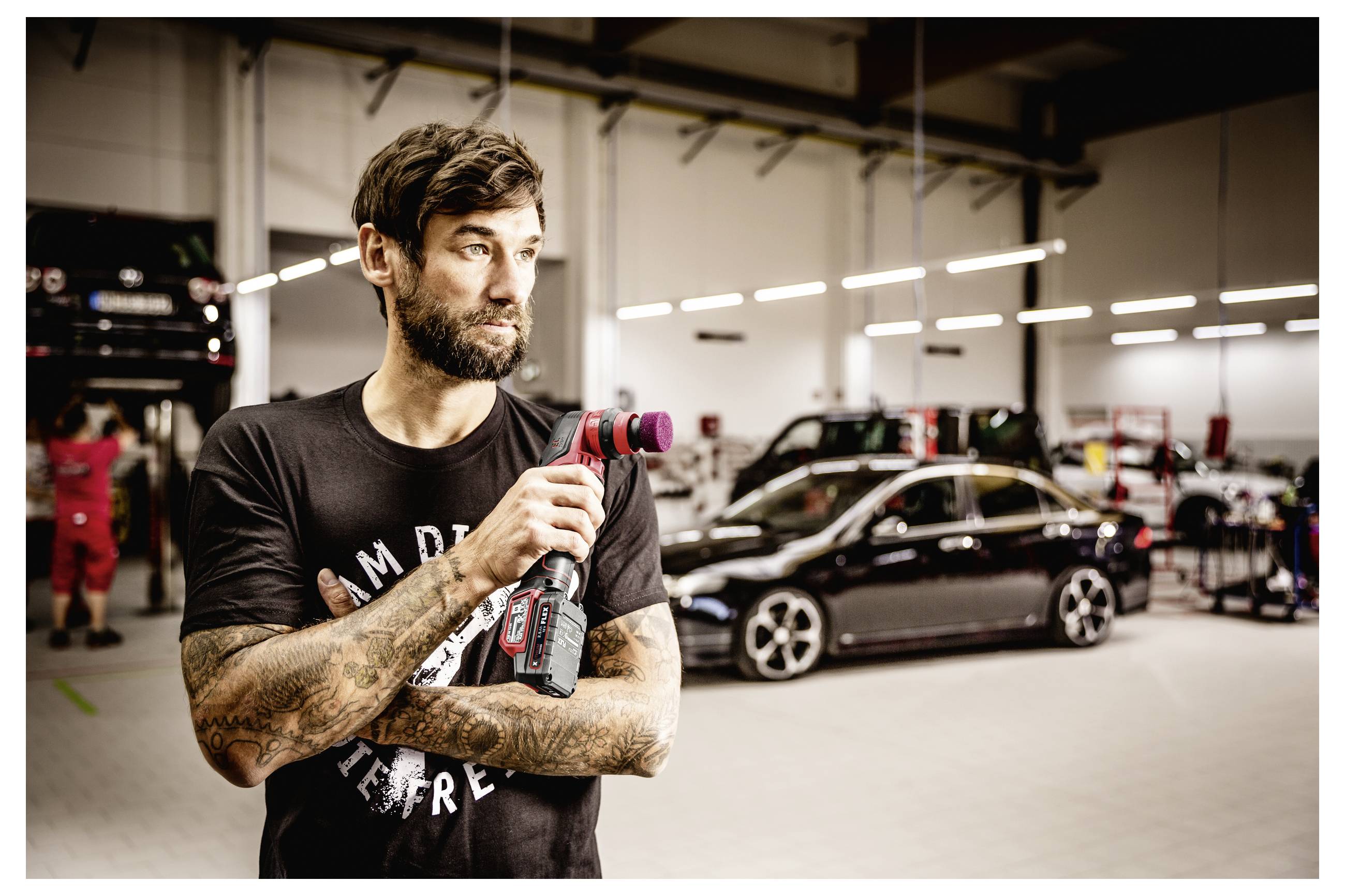 Tätowierter Mechaniker hält ein kabelloses Werkzeug in einer Autowerkstatt, mit Fahrzeugen und Ausrüstung im Hintergrund.