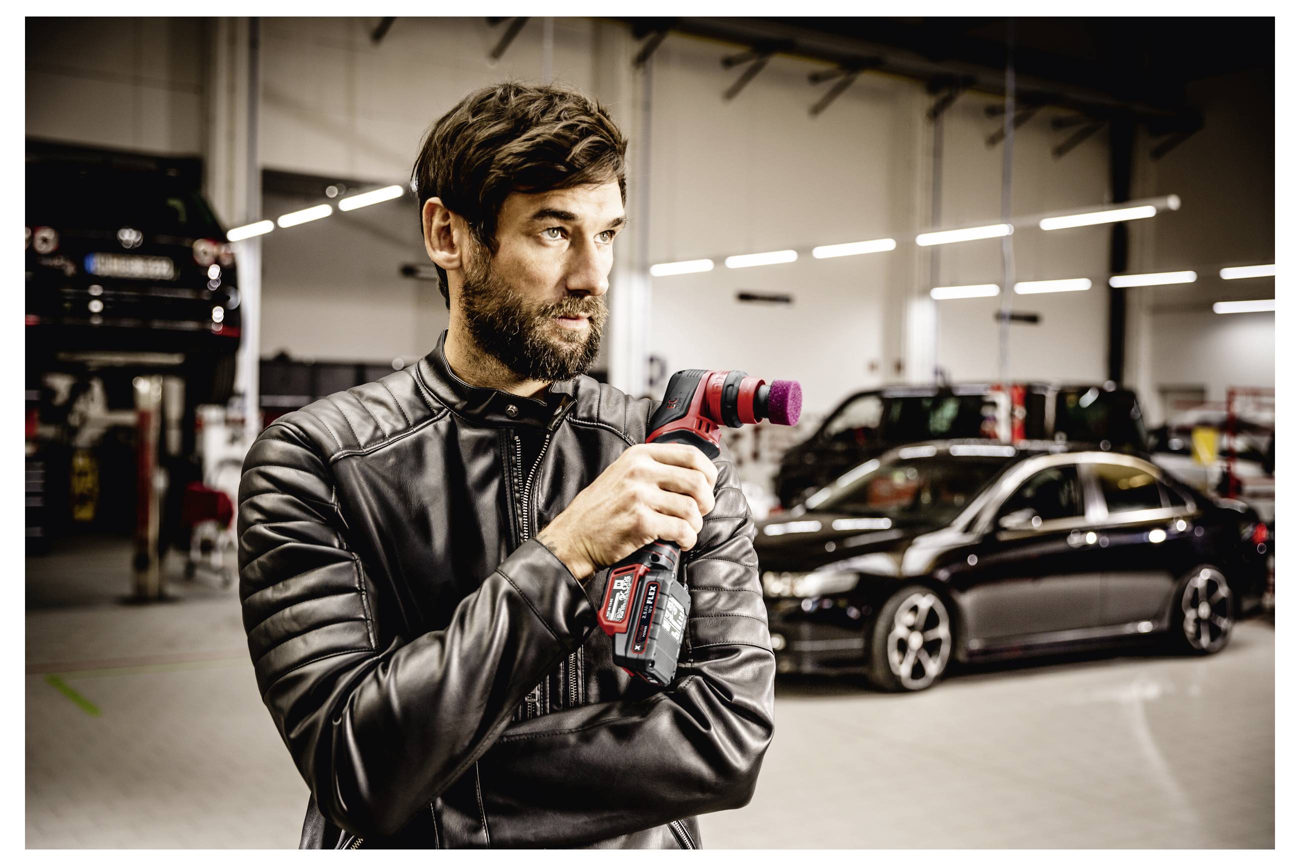 Ein Mann in einer schwarzen Lederjacke hält ein rotes Elektrowerkzeug in einer Autowerkstatt, mit Autos und einem aufgebockten Fahrzeug im Hintergrund.