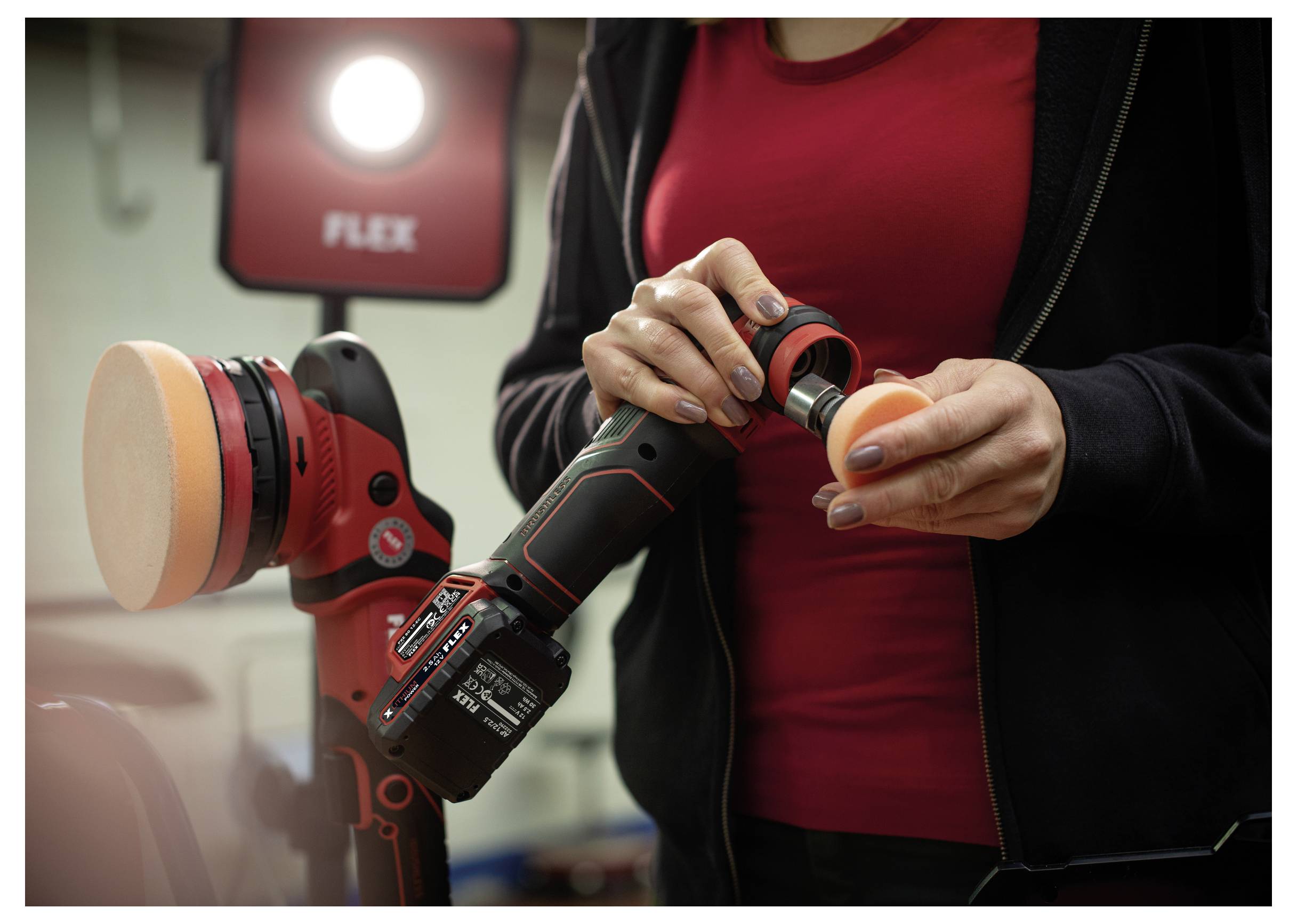 Person, die ein rotes schnurloses FLEX-Poliergerät einstellt, befestigt ein orangefarbenes Schaumstoffpad in einer Werkstatt-Ausstellungsumgebung.