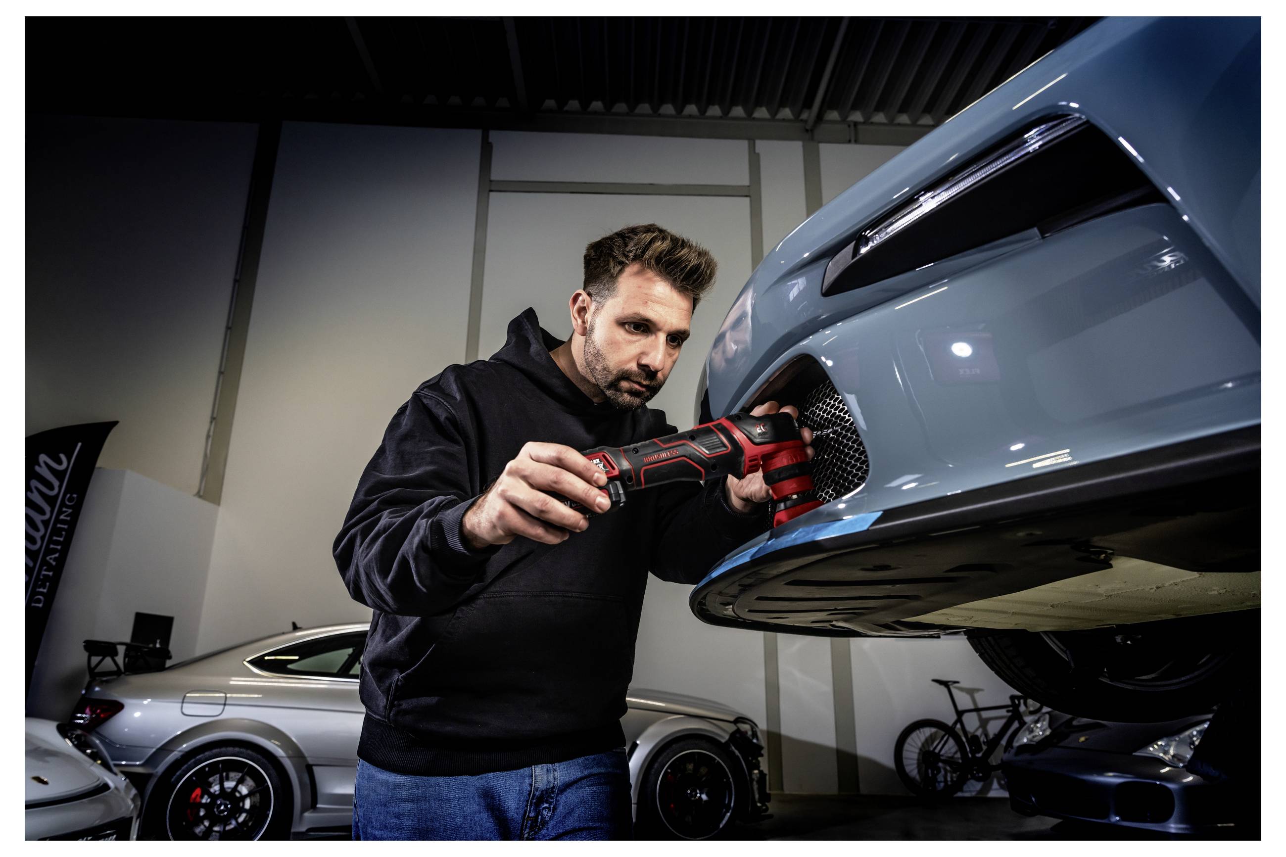 Ein Mechaniker verwendet ein Elektrowerkzeug, um an der Vorderseite eines blauen Sportwagens in einer Werkstatt zu arbeiten, mit anderen Autos im Hintergrund sichtbar.