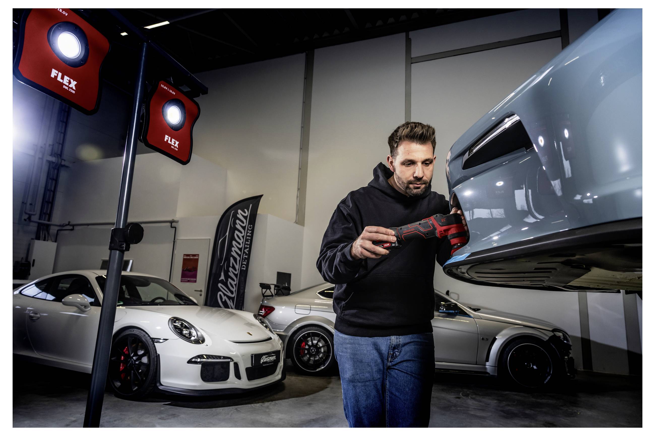 Ein Mann inspiziert und poliert die Vorderseite eines hellblauen Sportwagens in einer Garage, mit zwei weiteren Porsche-Autos und roten FLEX-Arbeitsleuchten im Hintergrund.