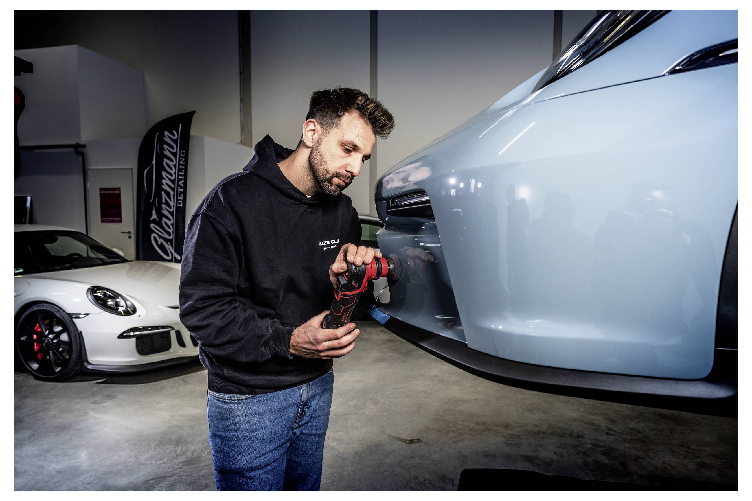 Ein Mechaniker benutzt ein Elektrowerkzeug, um die Seite eines hellblauen Sportwagens in einer Werkstatt zu polieren oder zu reparieren, während im Hintergrund ein anderes weißes Auto geparkt ist.