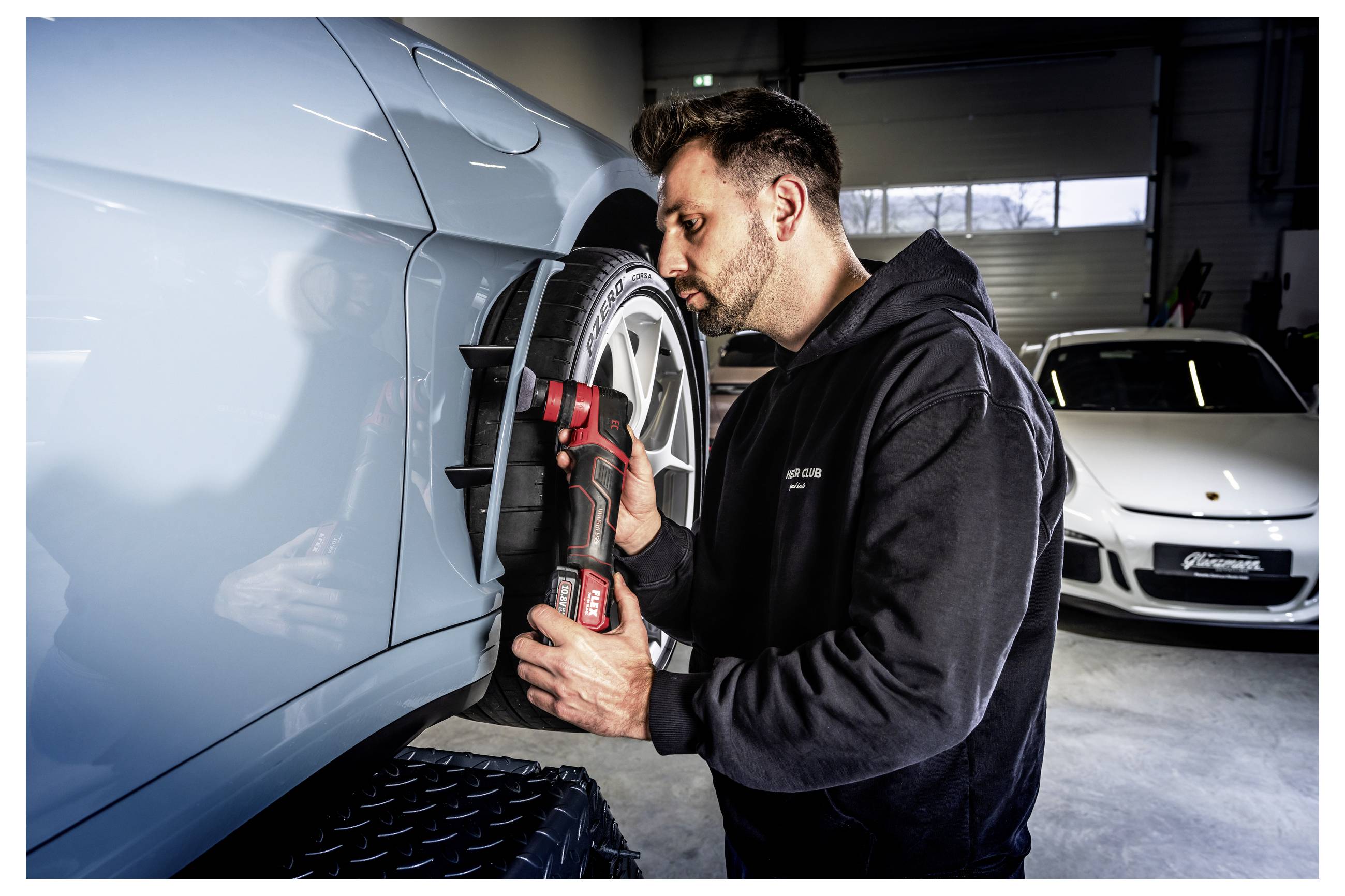 Ein Mechaniker inspiziert und poliert den Radbereich eines hellblauen Autos in einer Werkstatt, während ein weißer Sportwagen im Hintergrund geparkt ist.