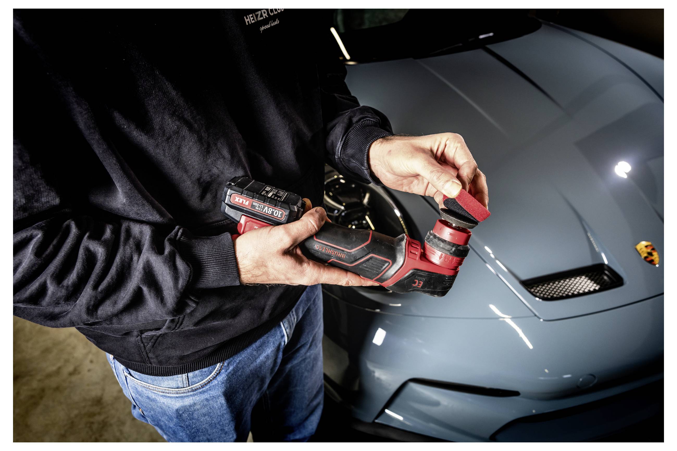Eine Person verwendet einen rot-schwarzen kabellosen Poliermaschine auf der Motorhaube eines blauen Sportwagens in einer Garage und verfeinert den Lackfinish.