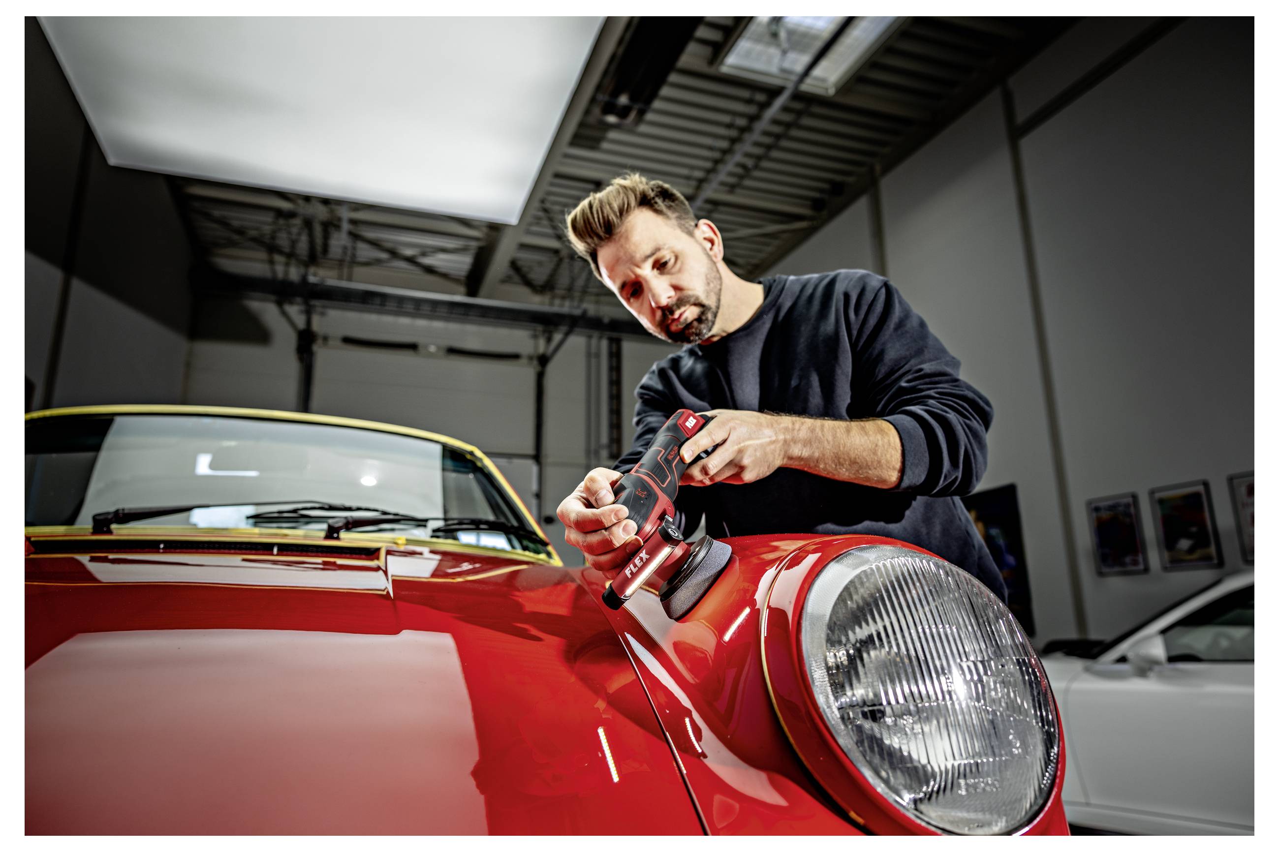 Mechaniker poliert die Motorhaube eines roten Oldtimers in einer Werkstatt und verwendet dabei einen tragbaren Poliergerät in der Nähe des vorderen Scheinwerfers.