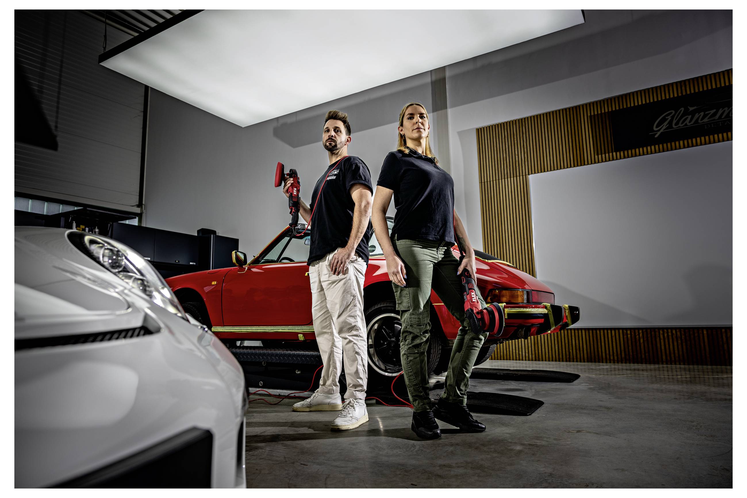 Zwei Mechaniker stehen in einer Werkstatt neben einem roten Sportwagen, mit einem weißen Auto im Vordergrund und einem Wandschild, auf dem „Glanz