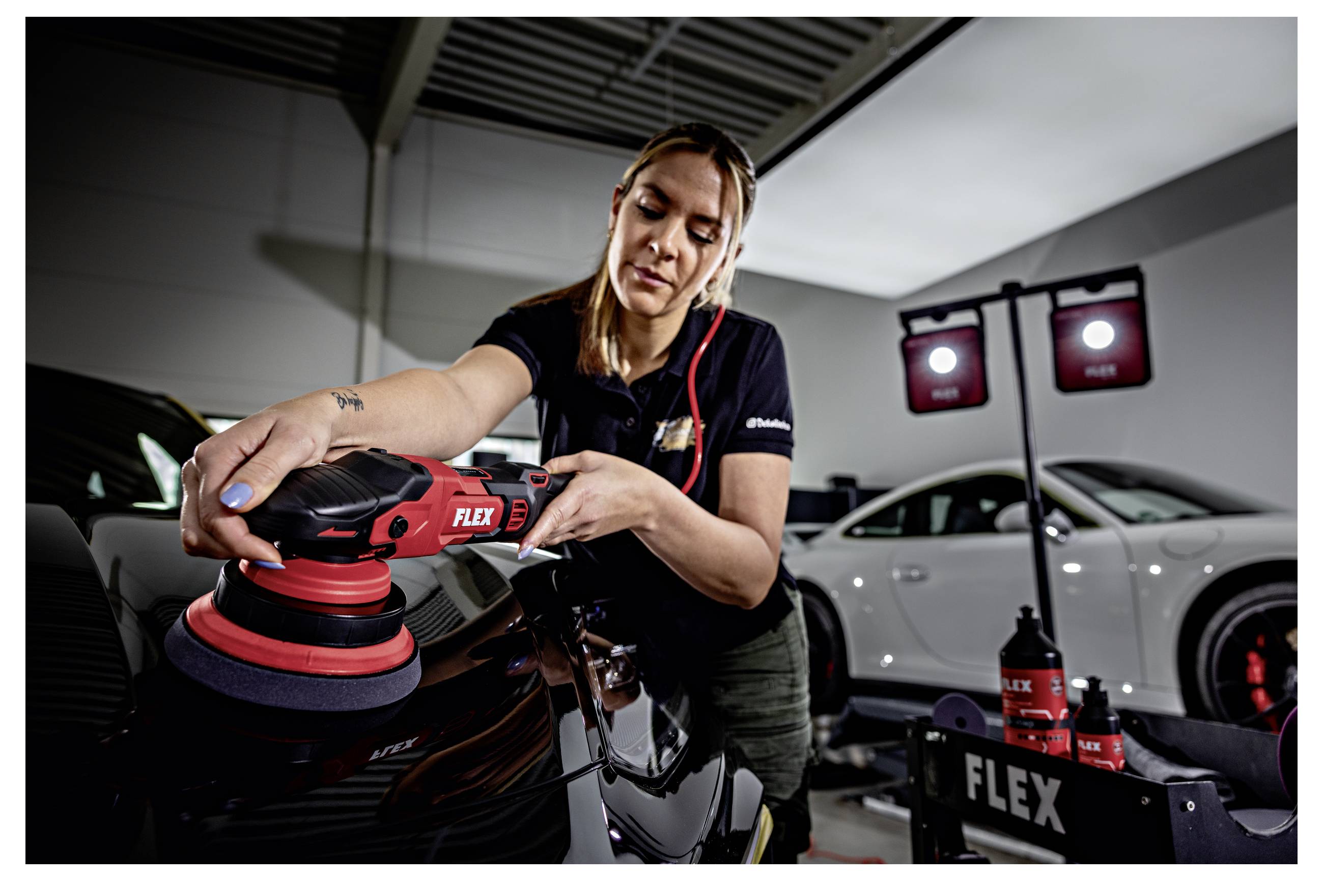 Techniker, der mit einem FLEX-Poliergerät ein Auto in einer Werkstatt poliert, mit einem weißen Sportwagen und Detailprodukten im Hintergrund.
