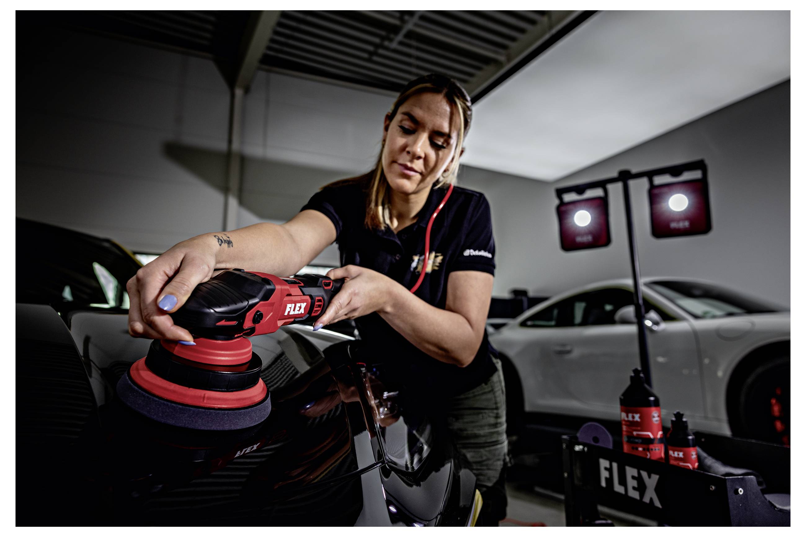 Frau, die ein schwarzes Auto mit einem roten FLEX-Poliergerät in einer Garage poliert, mit Detailing-Zubehör und einem weißen Auto im Hintergrund.