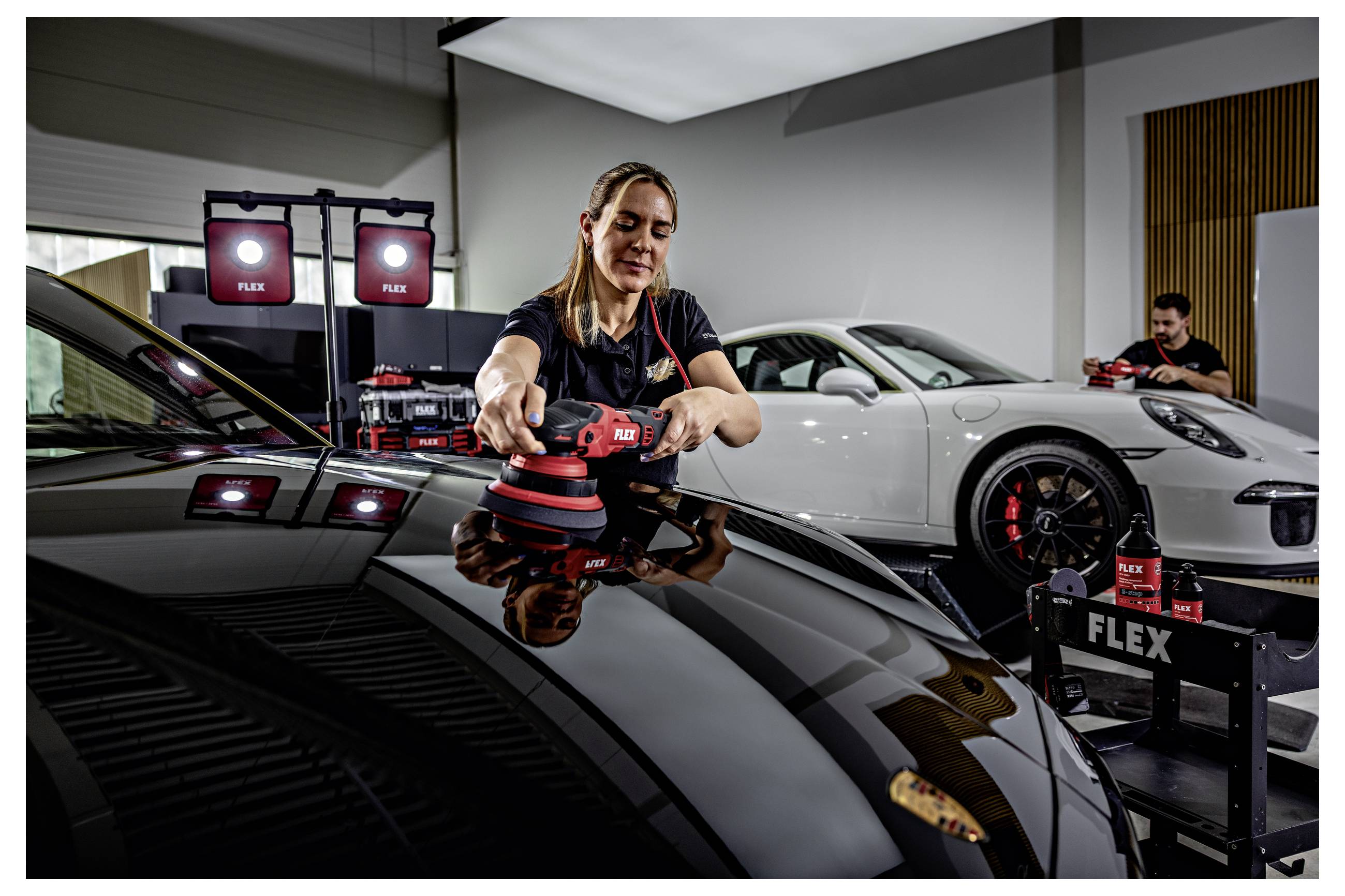 Ein Techniker poliert einen schwarzen Porsche mit einem FLEX-Poliergerät in einer Detaillierungswerkstatt, während ein weißer Porsche hinter ihm geparkt ist.