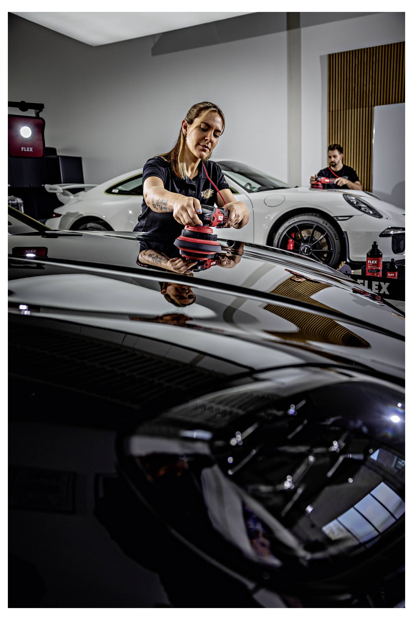 Ein Detailing-Techniker poliert die schwarze Motorhaube eines Autos mit einem Elektroschleiffer in einer Werkstatt, während ein weißer Sportwagen und ein weiterer Arbeiter im Hintergrund zu sehen sind.