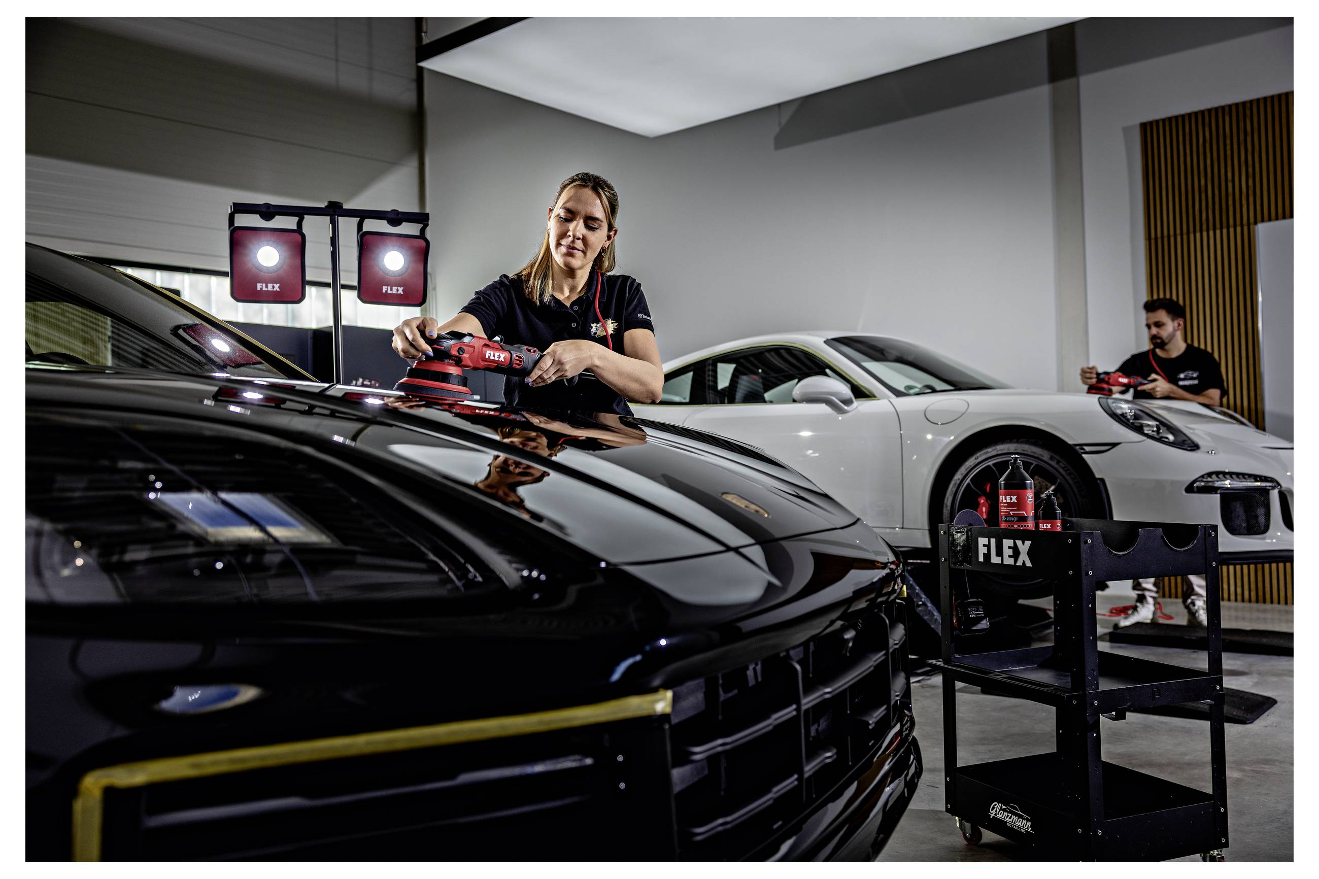 Ein Detailer poliert ein schwarzes Sportauto in einer Garage, mit einem weißen Porsche und tragbaren Leuchten im Hintergrund.