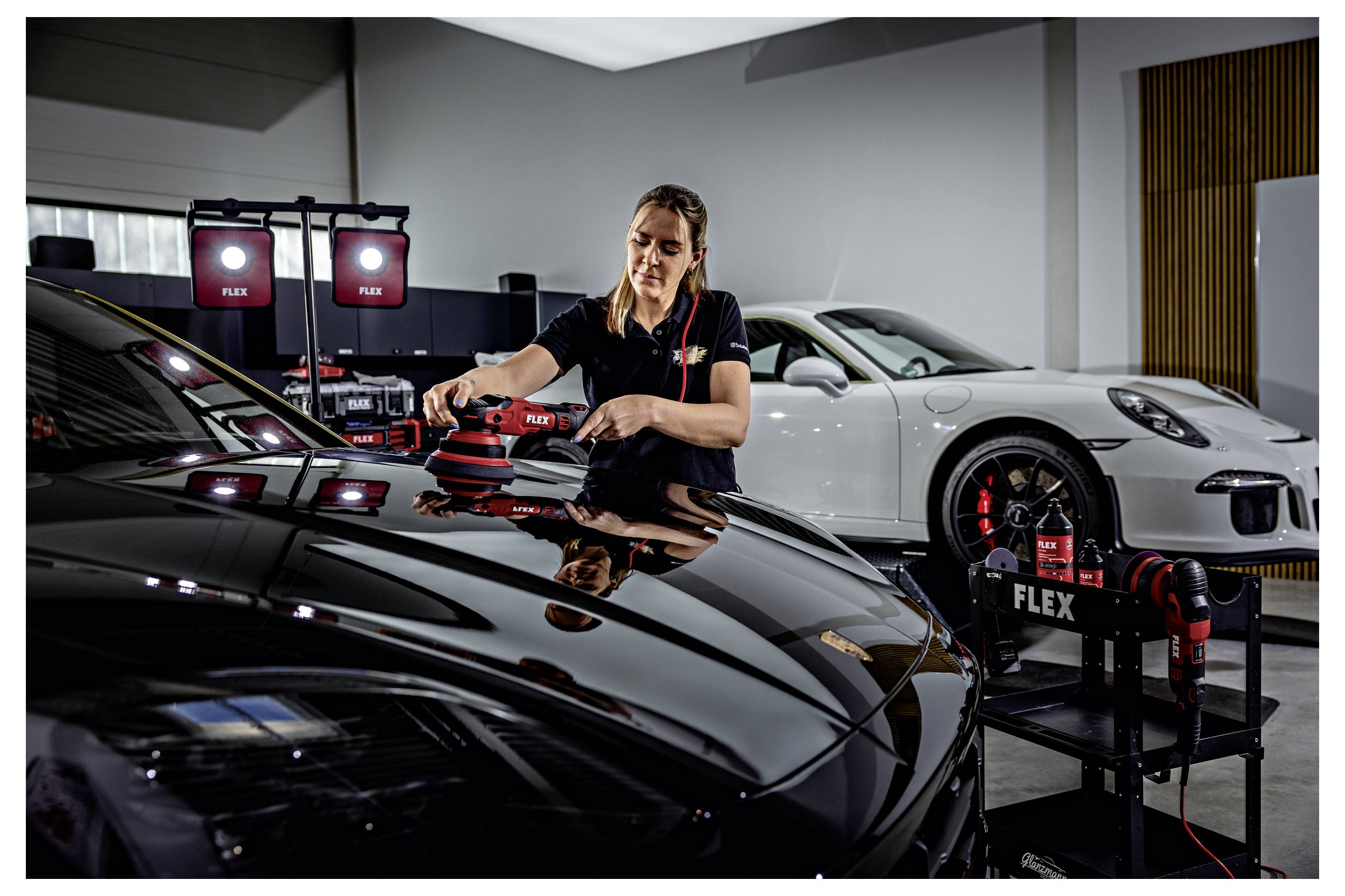 Ein Detailer poliert ein schwarzes Sportauto in einem Studio, mit Beleuchtungsausrüstung und einem weißen Sportauto im Hintergrund.
