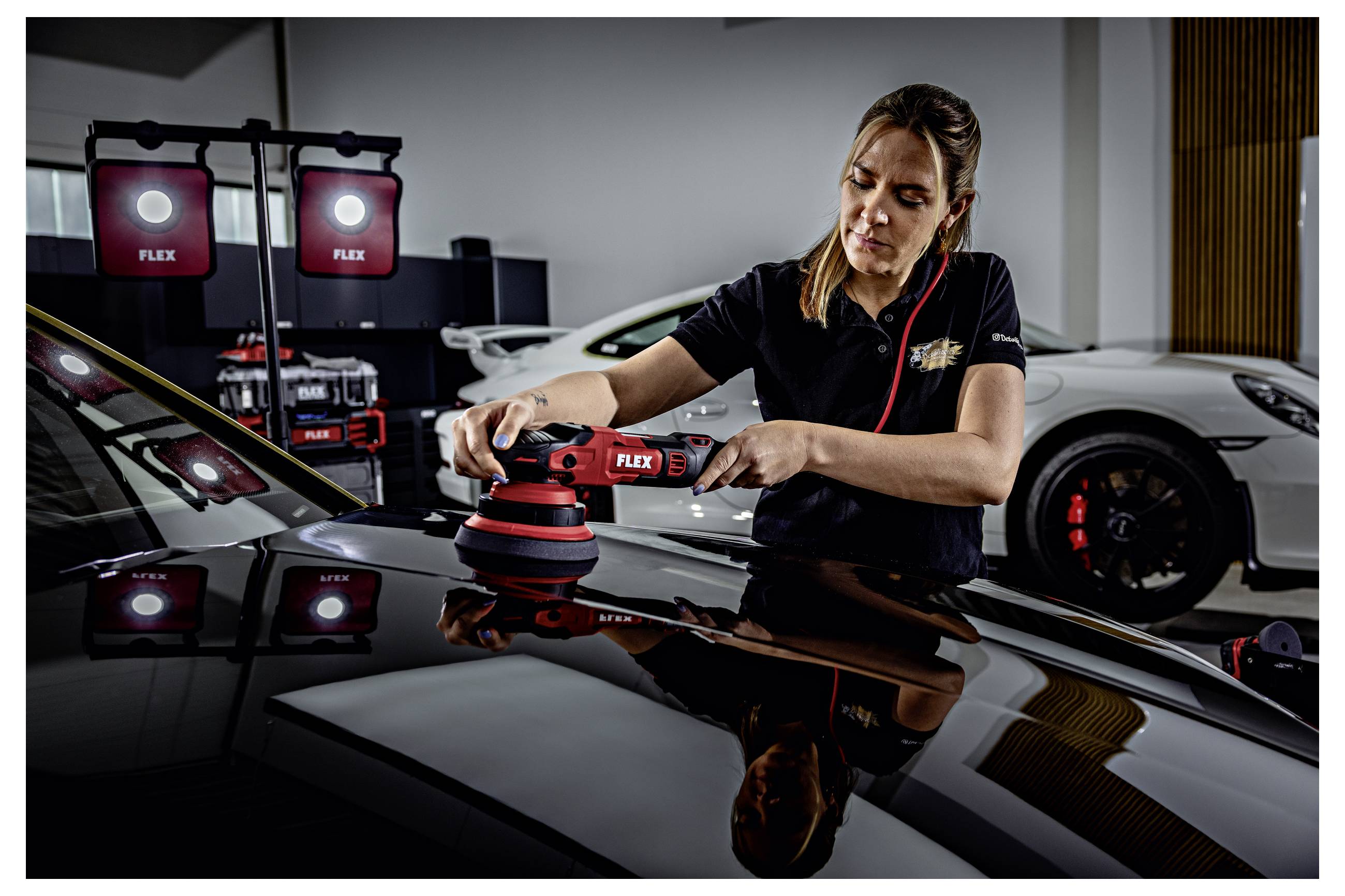 Techniker poliert eine Motorhaube eines Autos mit einem FLEX-Poliergerät in einer Detailing-Werkstatt, mit Studiolichtern und einem Sportwagen im Hintergrund.