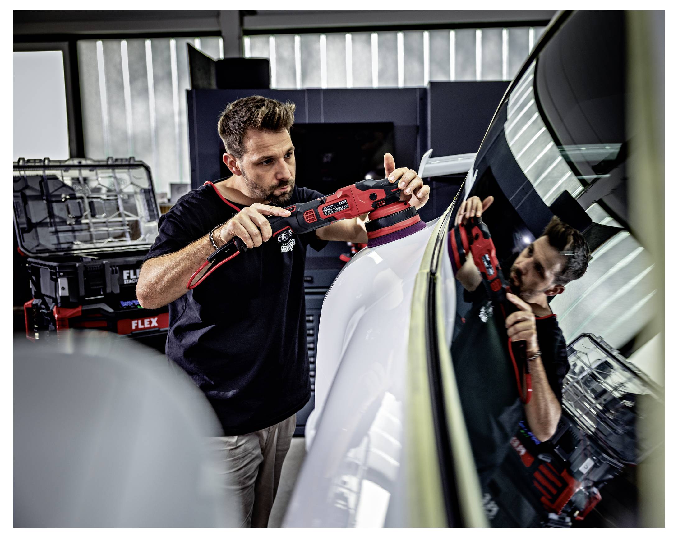 Ein Detaillierungstechniker poliert die Motorhaube eines Autos mit einem roten Elektroschleiffer in einer Werkstatt, wobei das Werkzeug und sein Spiegelbild in der Windschutzscheibe sichtbar sind.