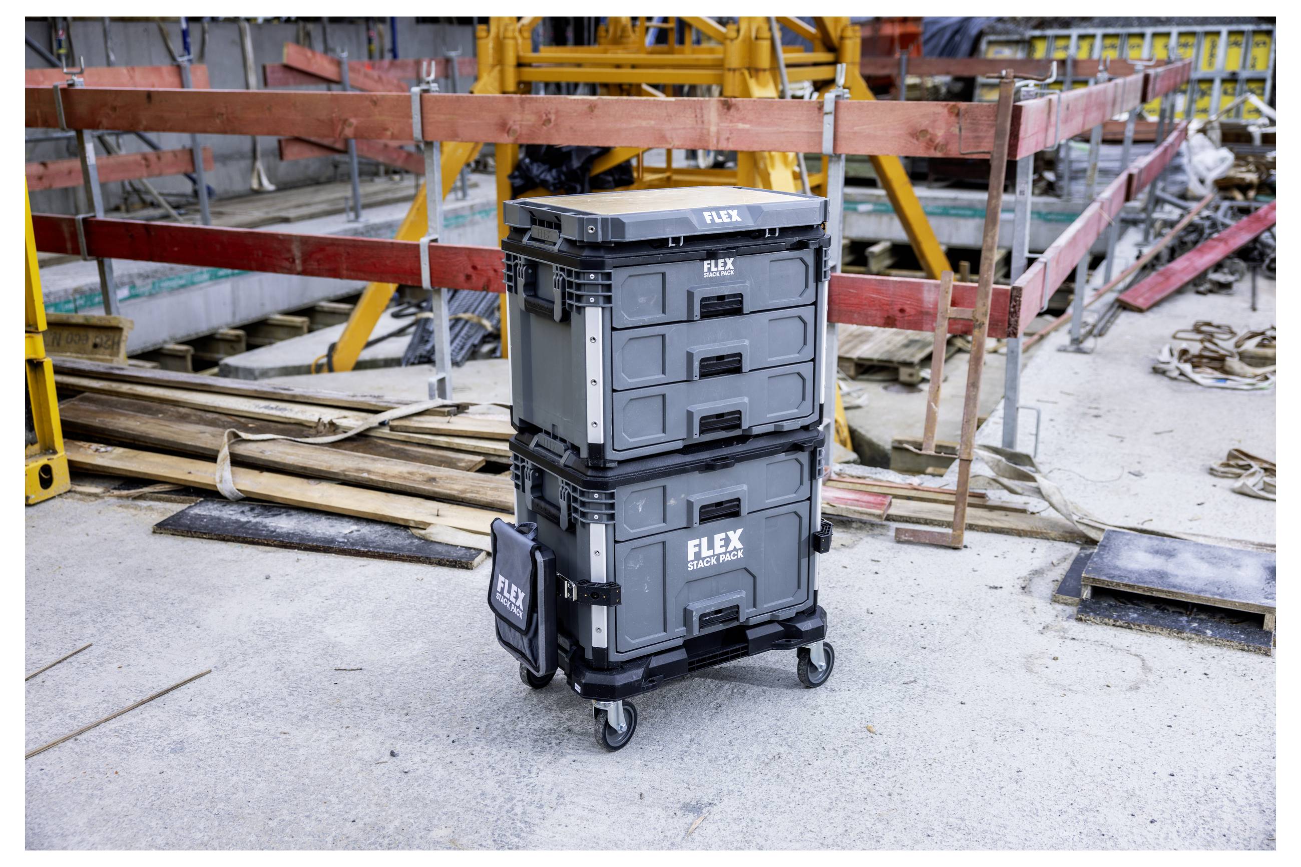Zwei gestapelte FLEX-Werkzeuglagerkästen auf einem Rollwagen auf einer Baustelle, mit Holz, Schienen und Ausrüstung im Hintergrund.