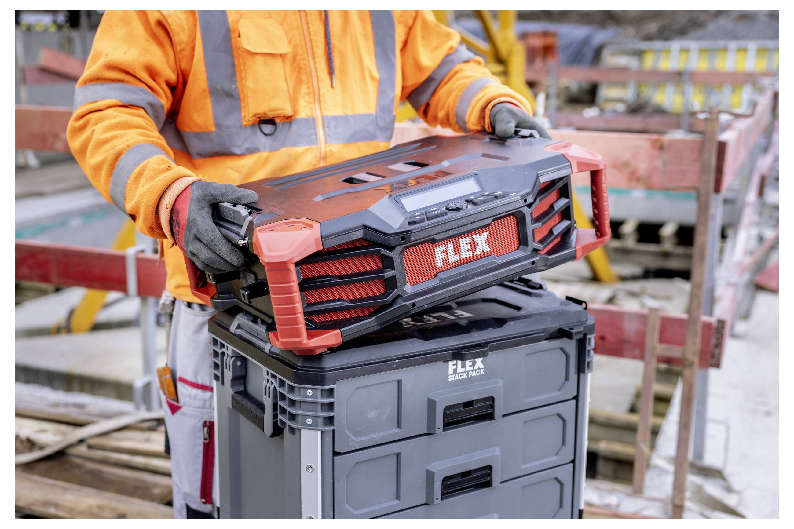 Bauarbeiter in orangefarbener Schutzausrüstung hebt eine tragbare FLEX-Werkzeugkiste von einer grauen Lagertruhe auf einer Baustelle.