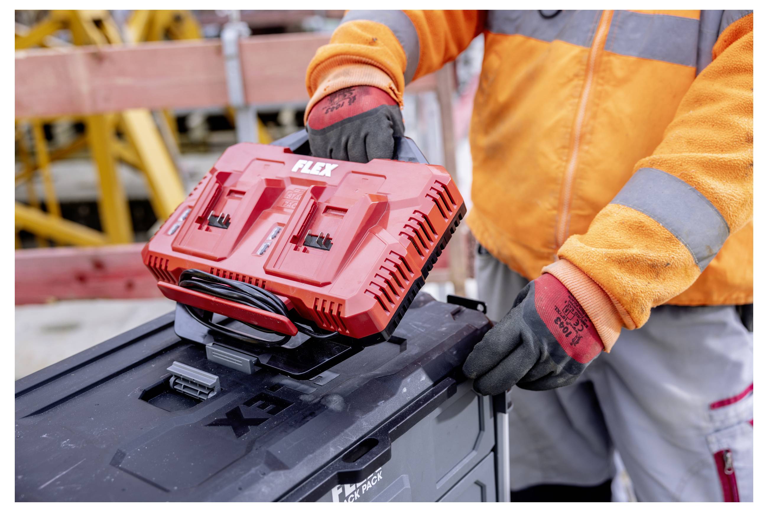 Arbeiter in orangefarbener Schutzausrüstung, der ein rotes FLEX-Werkzeug-Batterieladegerät auf einer schwarzen Werkzeugkiste auf einer Baustelle abstellt.