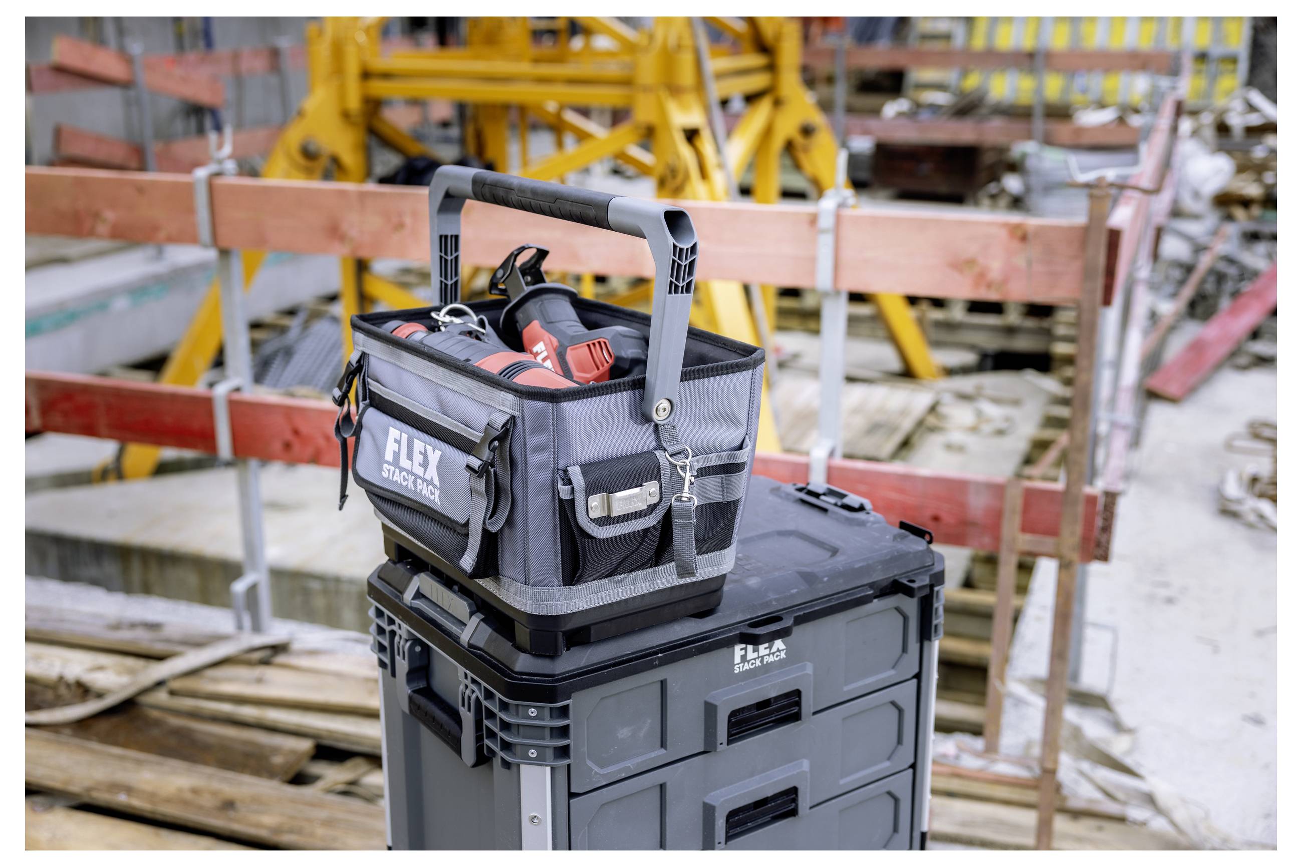 Werkzeugtasche mit Elektrowerkzeugen sitzt auf einem schwarzen Lagerbehälter auf einer Baustelle, umgeben von Gerüsten und Holzbarrieren.