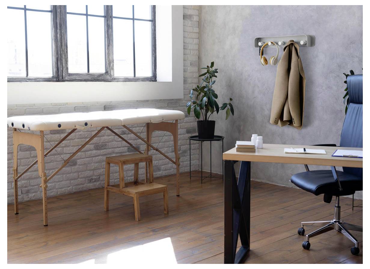 Minimalistische Therapiepraxis mit Massagetisch, Hocker, Schreibtisch, Stuhl, Garderobe und Pflanze in einem hellen Raum mit großen Fenstern.