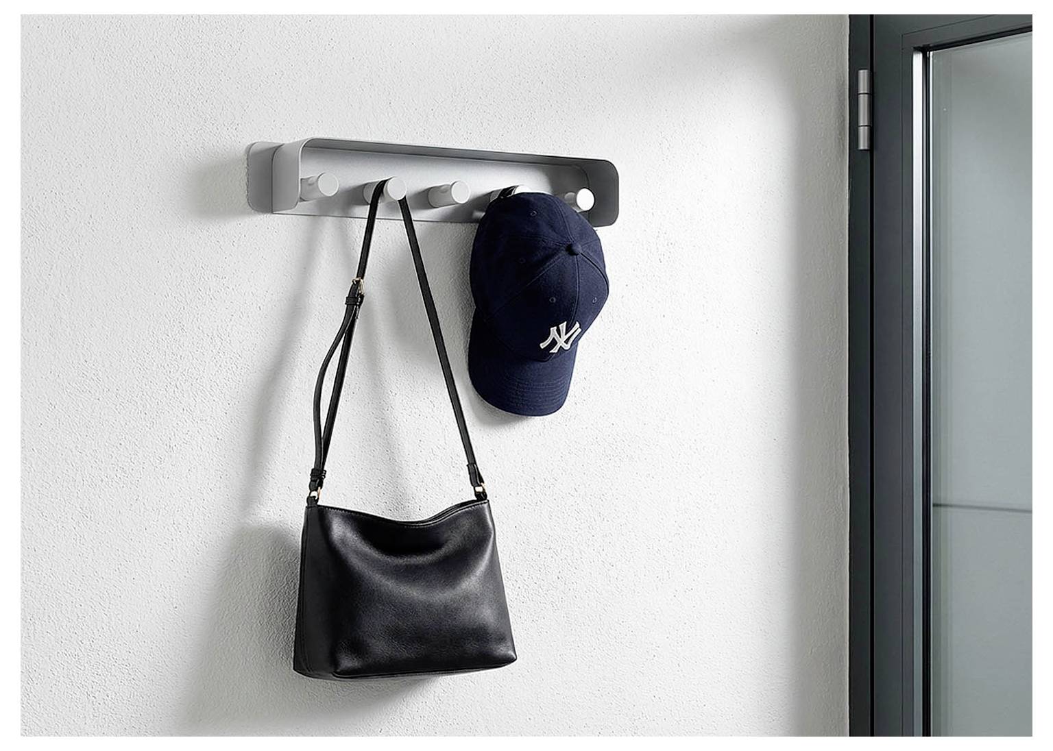 Wandmontierte Garderobe mit einer schwarzen Handtasche und einer dunkelblauen Baseballmütze, die an einer weißen Wand neben einer Tür hängen.