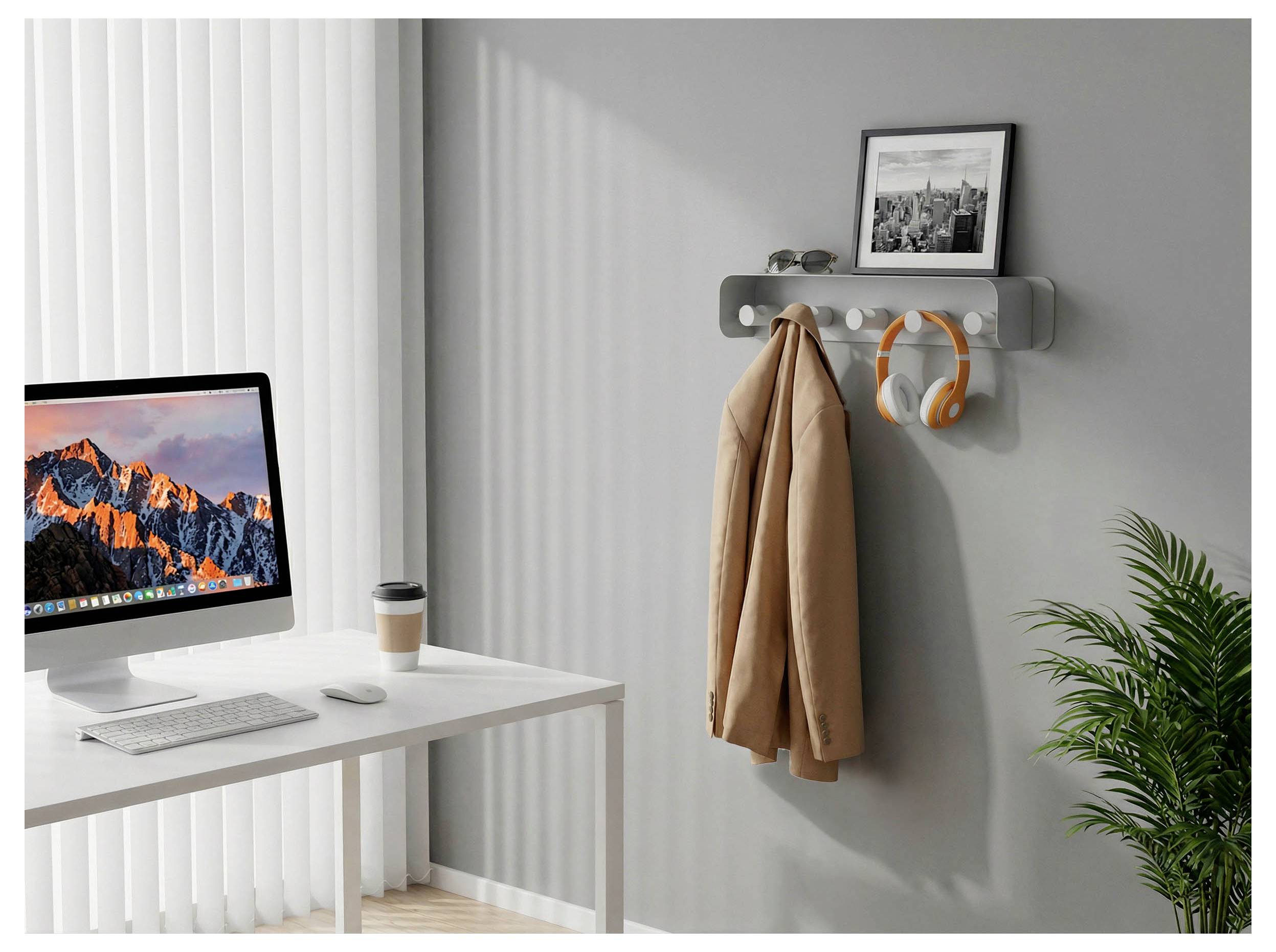 Minimales Homeoffice mit Schreibtisch, Desktop-Computer, Kaffeetasse, Wandhaken, an denen ein Mantel und Kopfhörer hängen, gerahmtem Foto und Topfpflanze.