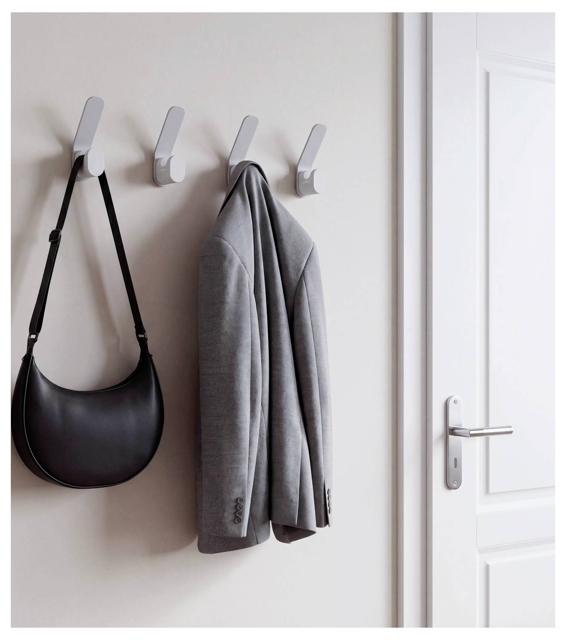 An der Wand befestigte Haken halten eine schwarze Schultasche und eine graue Jacke neben einer weißen Tür.