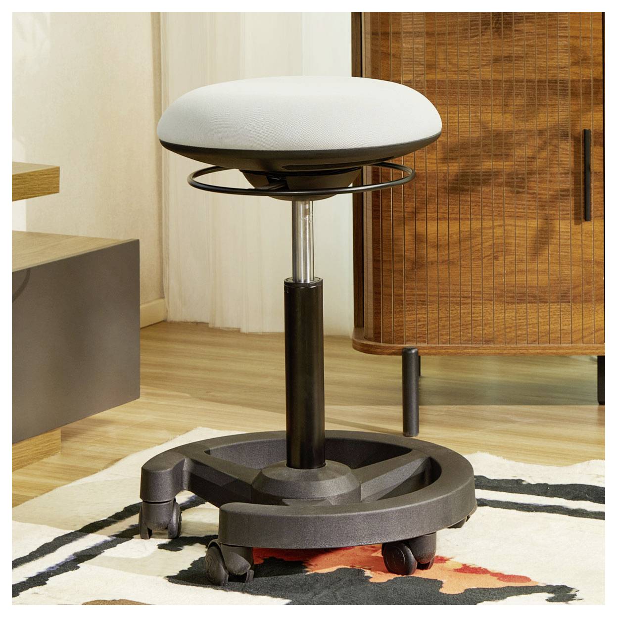 Verstellbarer Drehhocker mit gepolstertem weißem Sitz, schwarzem Fuß und Rollen auf einem gemusterten Teppich in einem modernen Zimmer.