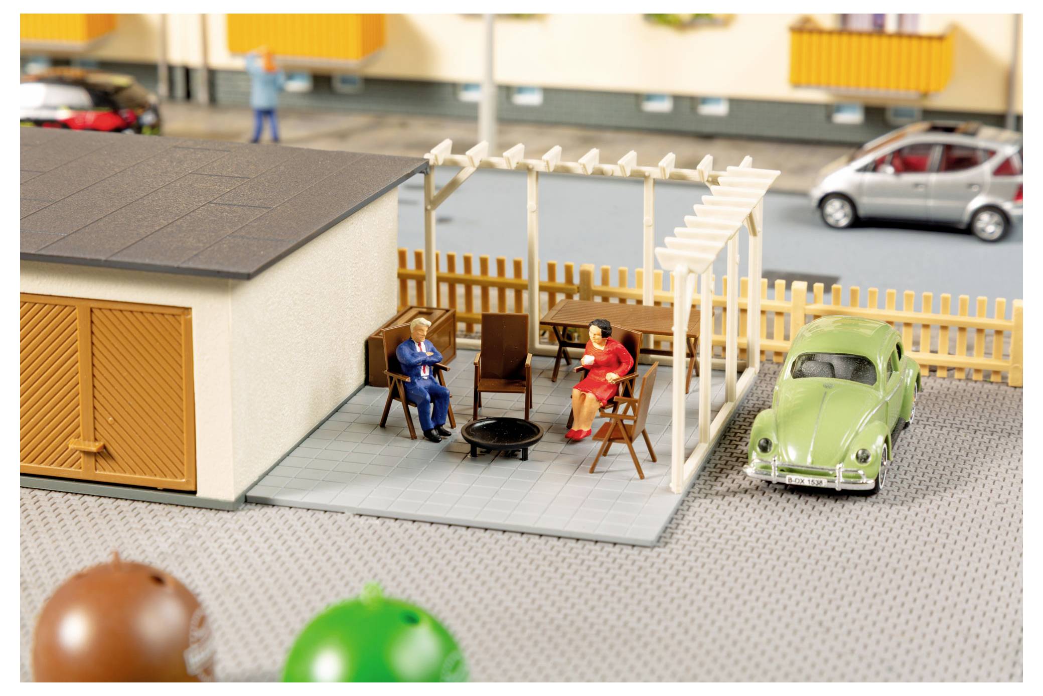 Miniatur-Hofszene mit zwei sitzenden Figuren, einem kleinen Tisch und Stühlen neben einer Garage, mit einem grünen Oldtimer-Auto in der Nähe geparkt.