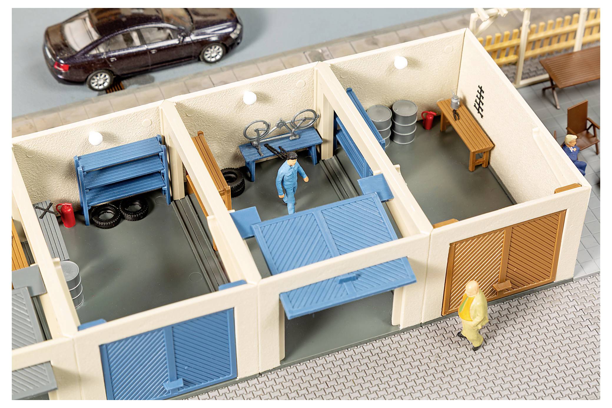 Spielzeug-Draufsicht eines Lagerhaus-/Werkstatt-Dioramas mit blauen Garagentoren, Arbeitern, gestapelten Fässern, einem Fahrrad und geparkten Autos draußen.