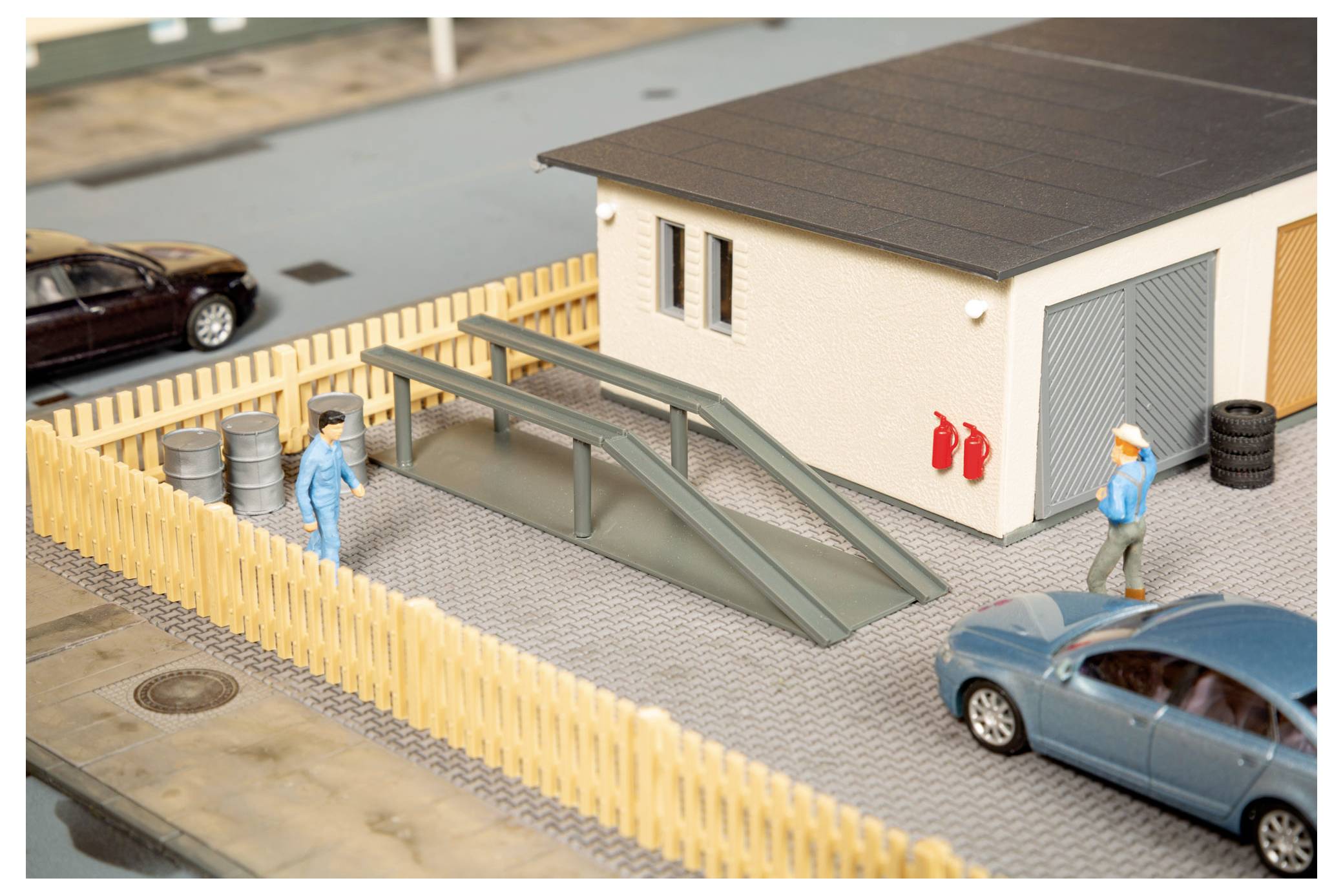 Miniatur-Straßenszene mit einem cremefarbenen Gebäude, eingezäuntem Grundstück, Rampenstruktur, zwei Arbeiterfiguren, geparkten Autos und roten Feuerlöschern.