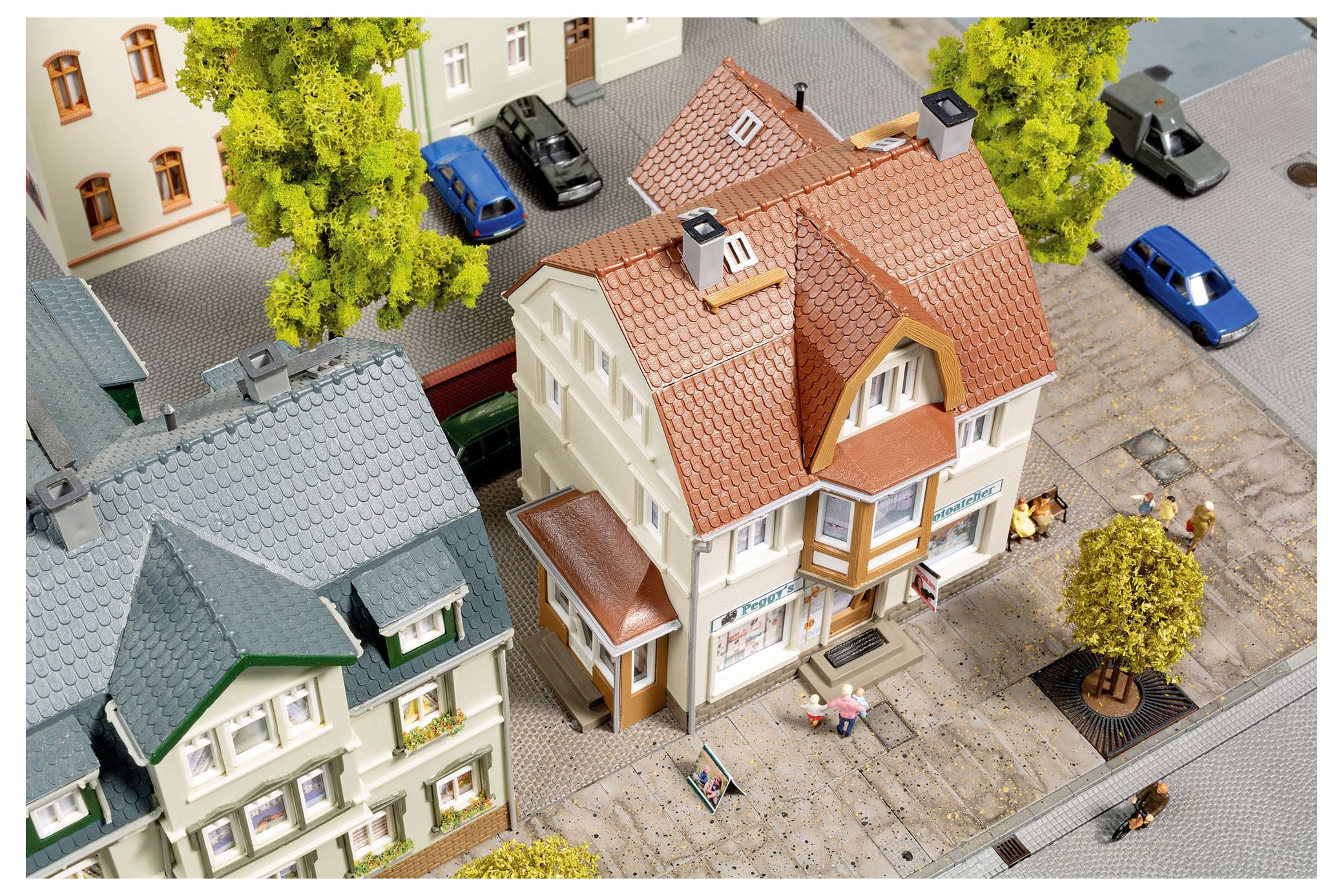 Miniaturstraßenszene mit Modellhäusern, geparkten Autos, Bäumen und winzigen Figuren auf einem Gehweg, von oben betrachtet.