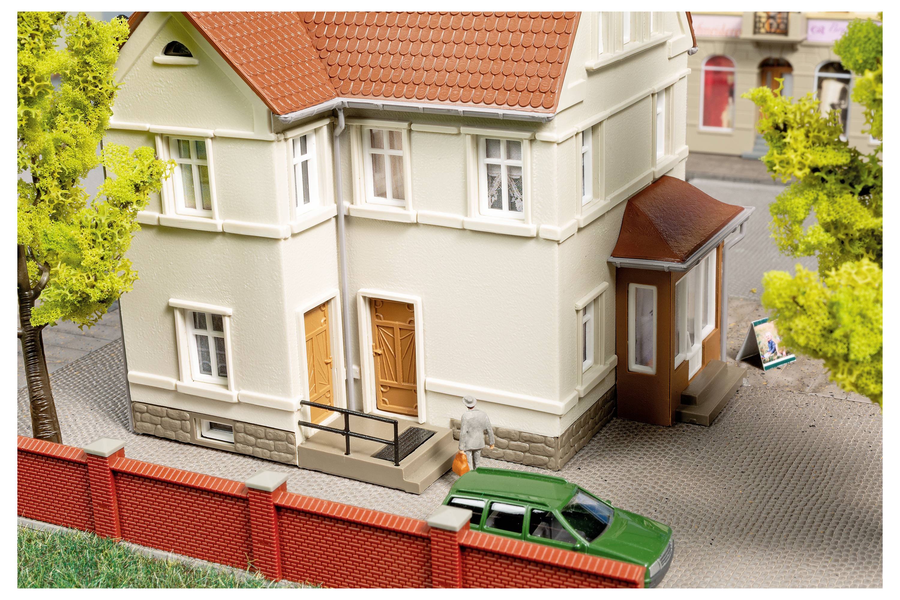 Modell eines cremefarbenen zweistöckigen Hauses mit orangefarbenem Dach, roter Backsteinmauer und grünem Auto, das draußen geparkt ist.