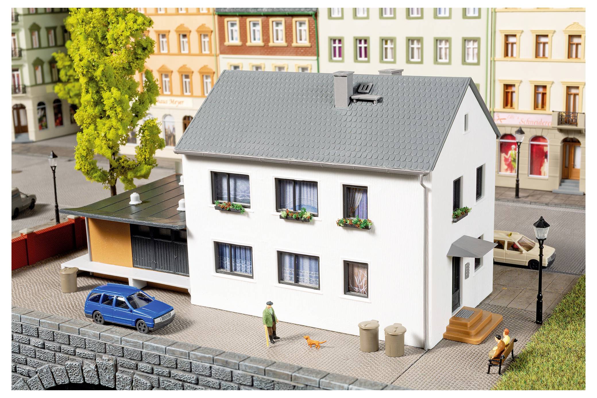 Modellstadtszene mit einem weißen zweistöckigen Haus neben einer Straße, blauem Auto, Fußgängern, Bänken, Bäumen und bunten Wohngebäuden im Hintergrund.