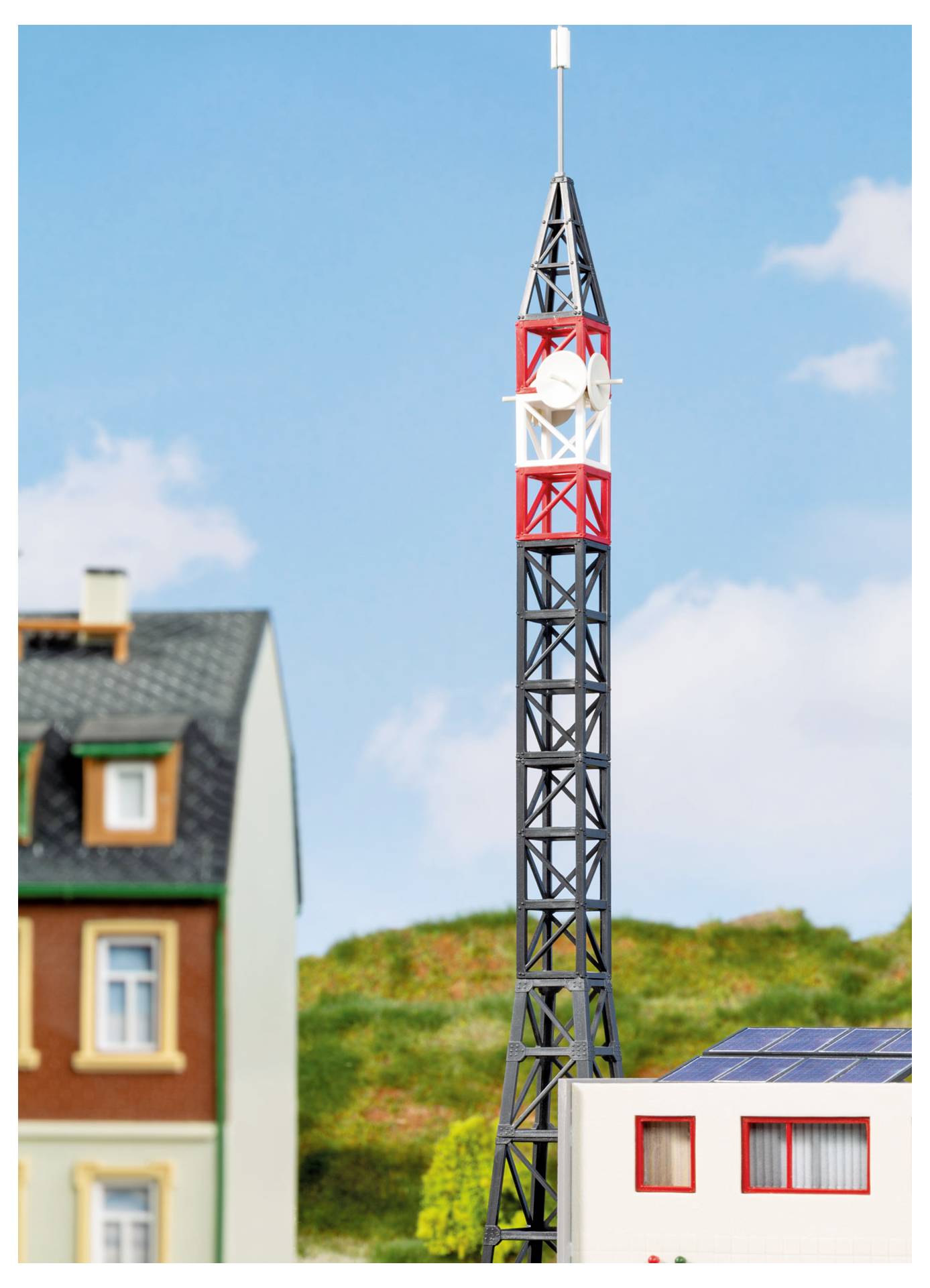Modellturm mit rot-weiß gestreifter Spitze steht neben Miniaturbäusern und einem Dach mit Solarpanelen unter blauem Himmel, was ein Maßstabsmodell einer Siedlung oder Telekommunikationsanlage andeutet.
