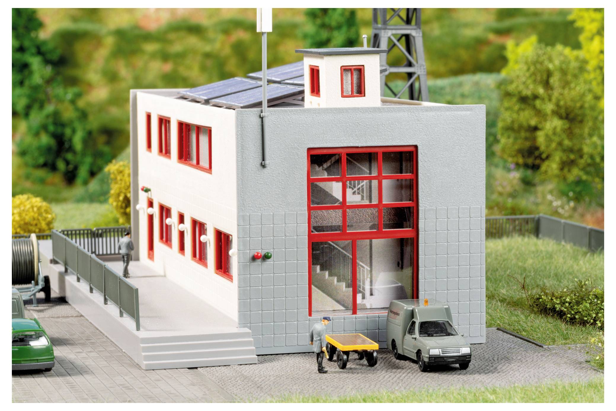 Miniatur-Industriegebäudemodell mit rot eingefassten Fenstern, Solarpanelen auf dem Dach, Treppen und kleinen Fahrzeugen außen auf einer Modelllandschaft.