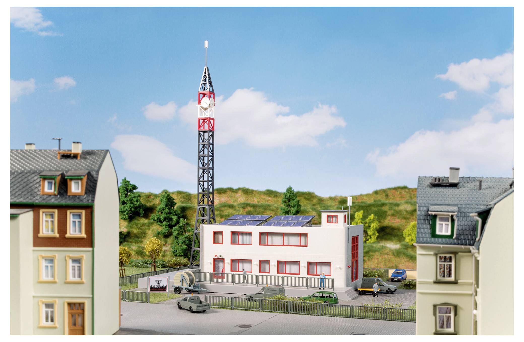 Modell eines kleinen Stadtblocks mit einem hohen Funkturm, einem zentralen modernen Gebäude mit Solarpanelen und umgebenden Häusern unter blauem Himmel.
