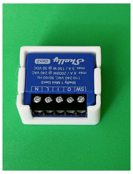 Blue Shelly 1 Mini Gen3 Smart-Relaismodul in einem weißen Halter auf grünem Hintergrund, zeigt Schraubklemmen und gedruckte Spezifikationen.