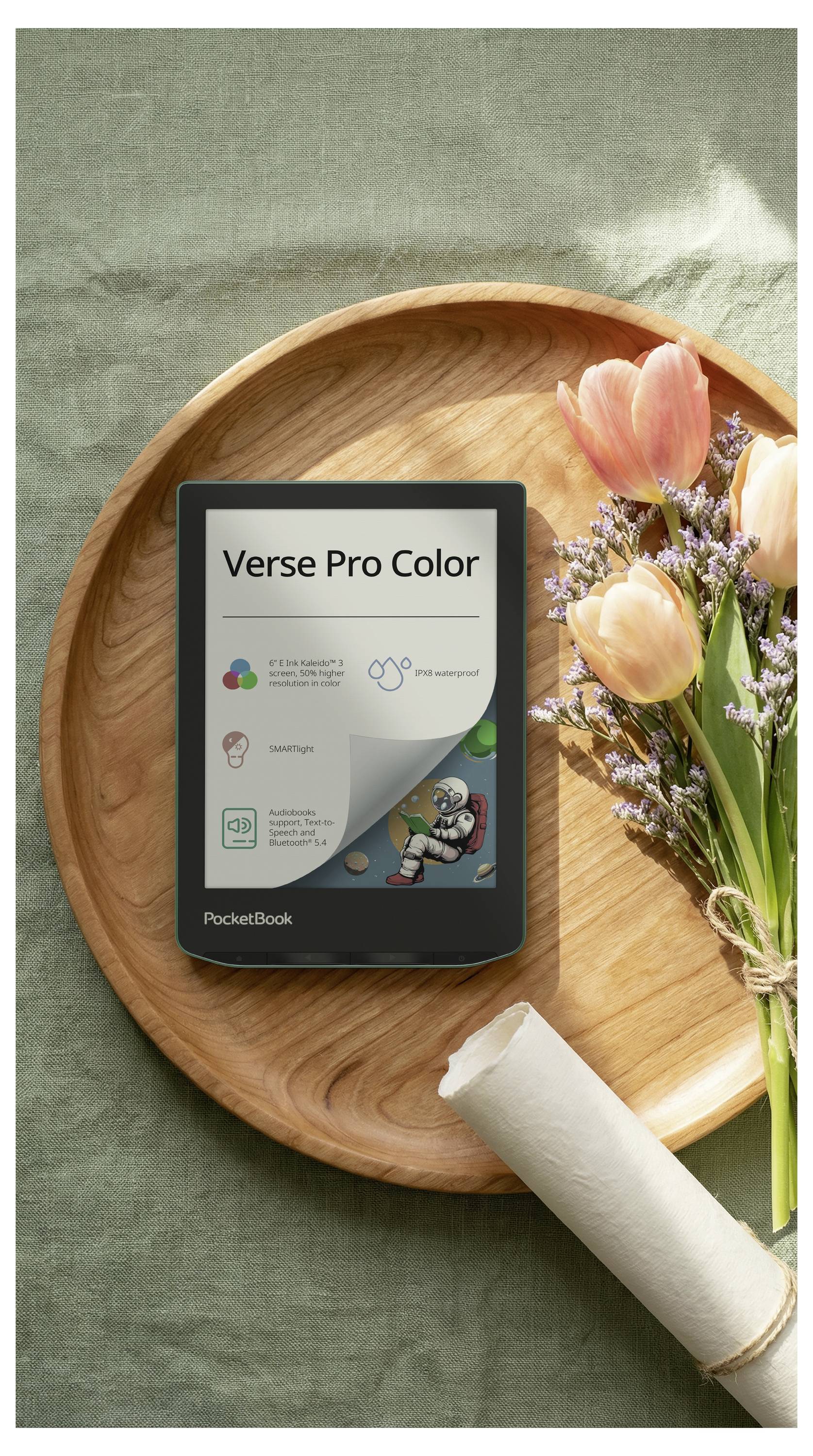 PocketBook Verse Pro Color E-Reader auf einem Holztablett neben Blumen und einer aufgerollten Serviette auf grünem Stoffhintergrund.