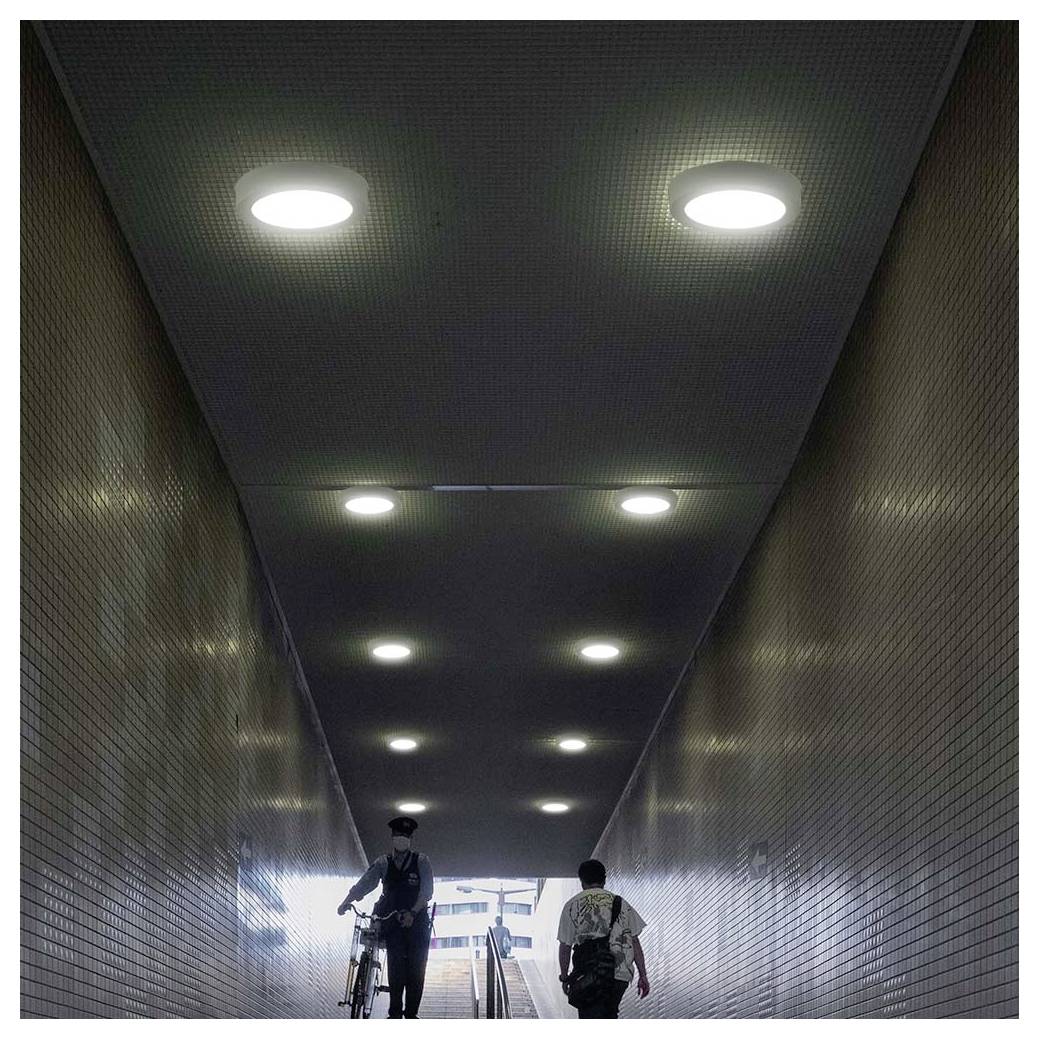 Tiefenwinkelperspektive durch einen gefliesten Tunnel mit Deckenleuchten, die einen Radfahrer und einen Fußgänger zeigt, die auf einen hellen Ausgang zugehen.