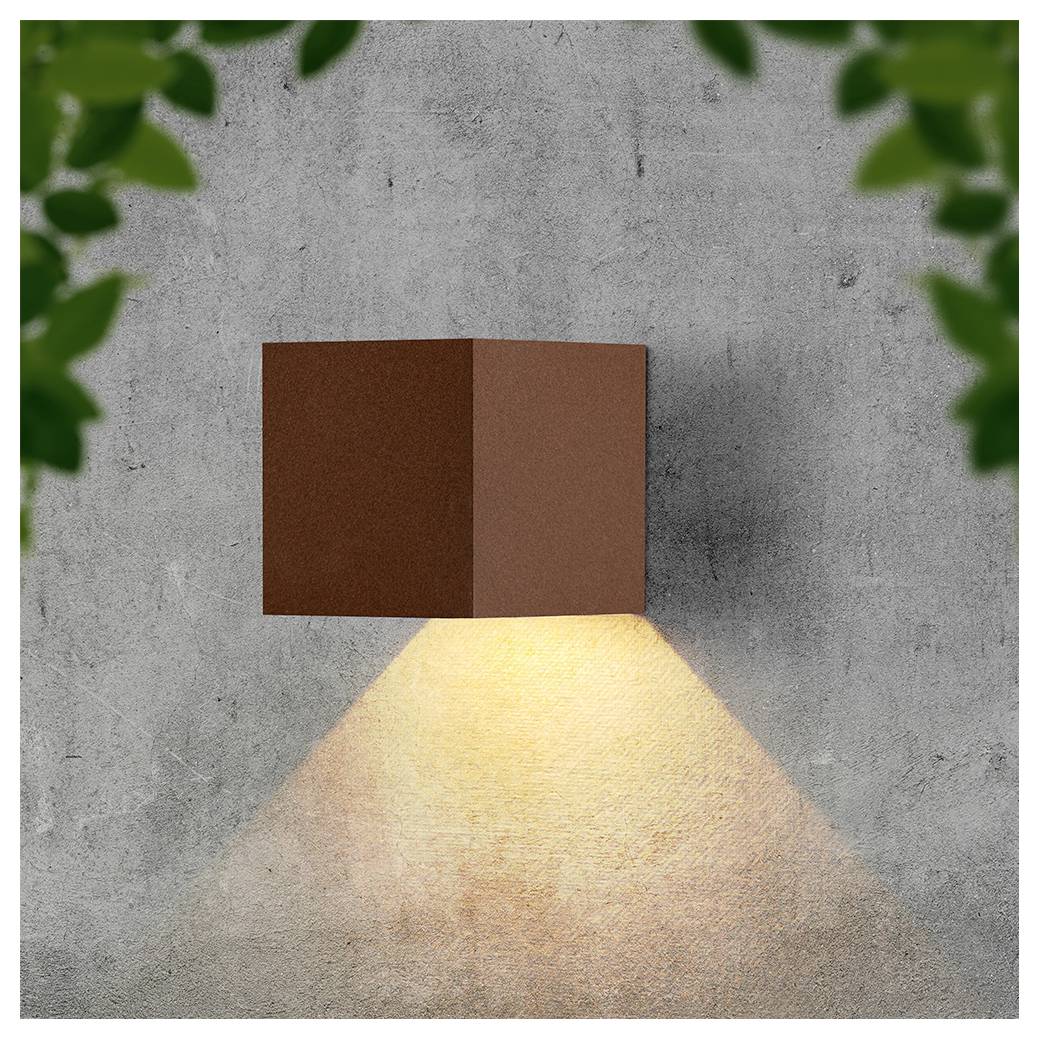 An der Wand montierte braune Würfelleuchte wirft einen warmen dreieckigen Lichtstrahl nach unten auf eine graue Betonwand, umrahmt von unscharfen grünen Blättern.