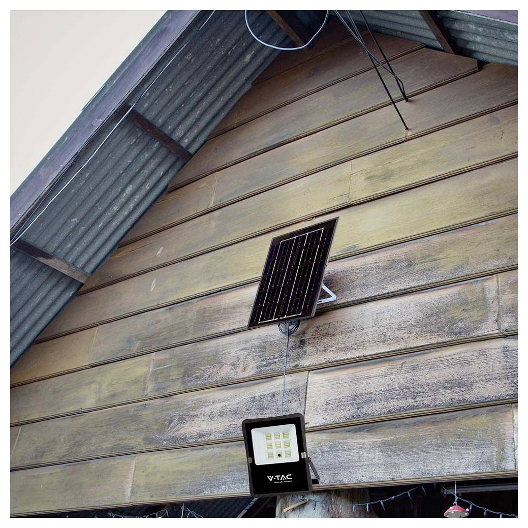 Ein kleines Solarpanel, das an einer Holzwand montiert ist, versorgt einen VTAC-LED-Flutlichtstrahler, der darunter unter einem gewellten Metalldach hängt.