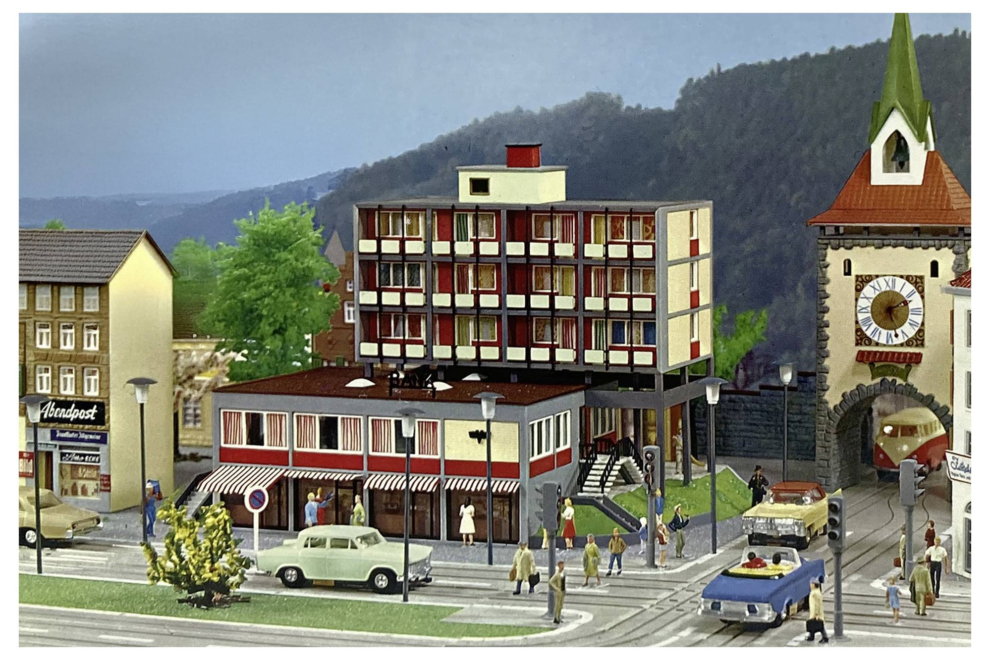 Modellhafte Straßenszene einer Stadt mit Fußgängern, Autos, einem zentralen Hotelgebäude und einem Torgebäude mit Uhr vor einer Bergkulisse.