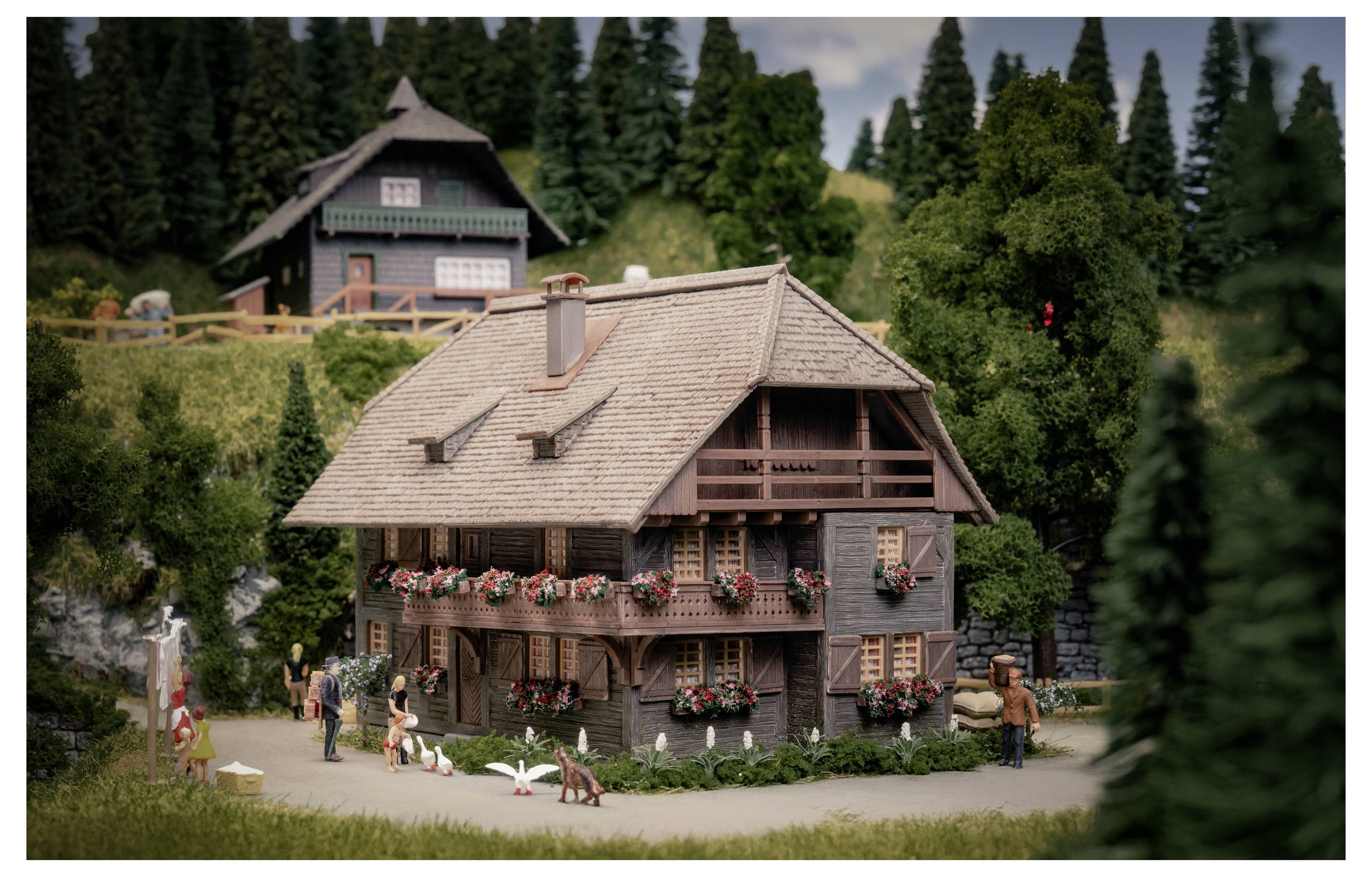 Miniaturmodell eines chaletförmigen Hauses mit Blumenkästen, kleinen Figuren und Schwänen in einer gestalteten Dorfszene mit Bäumen und einem weiteren Haus im Hintergrund.