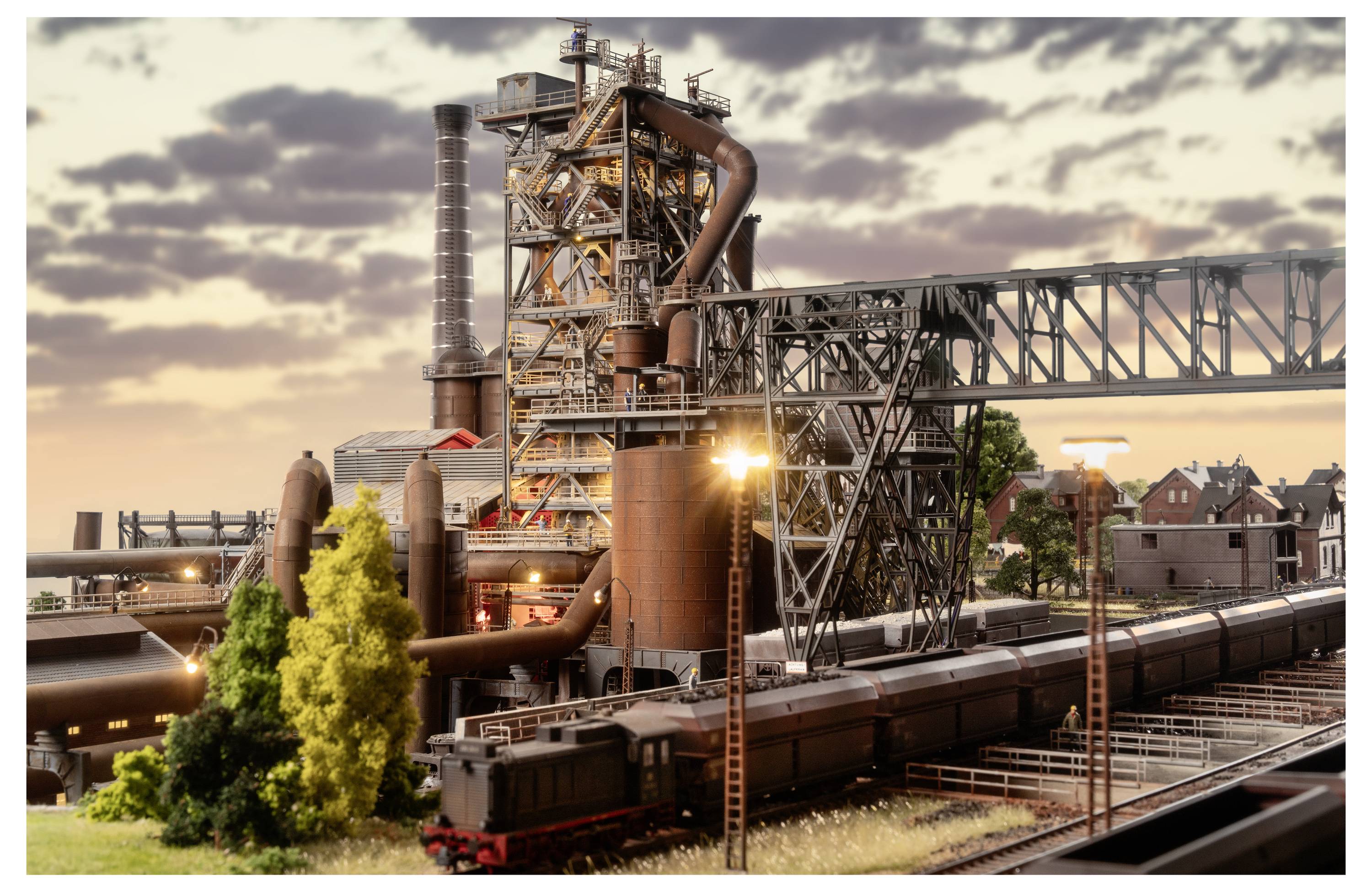 Stahlwerke in der Industrie mit Schornsteinen, Rohren und Eisenbahnwagen bei Sonnenuntergang, die einen großen Fabrikskomplex neben Wohngebäuden und Bäumen zeigen.