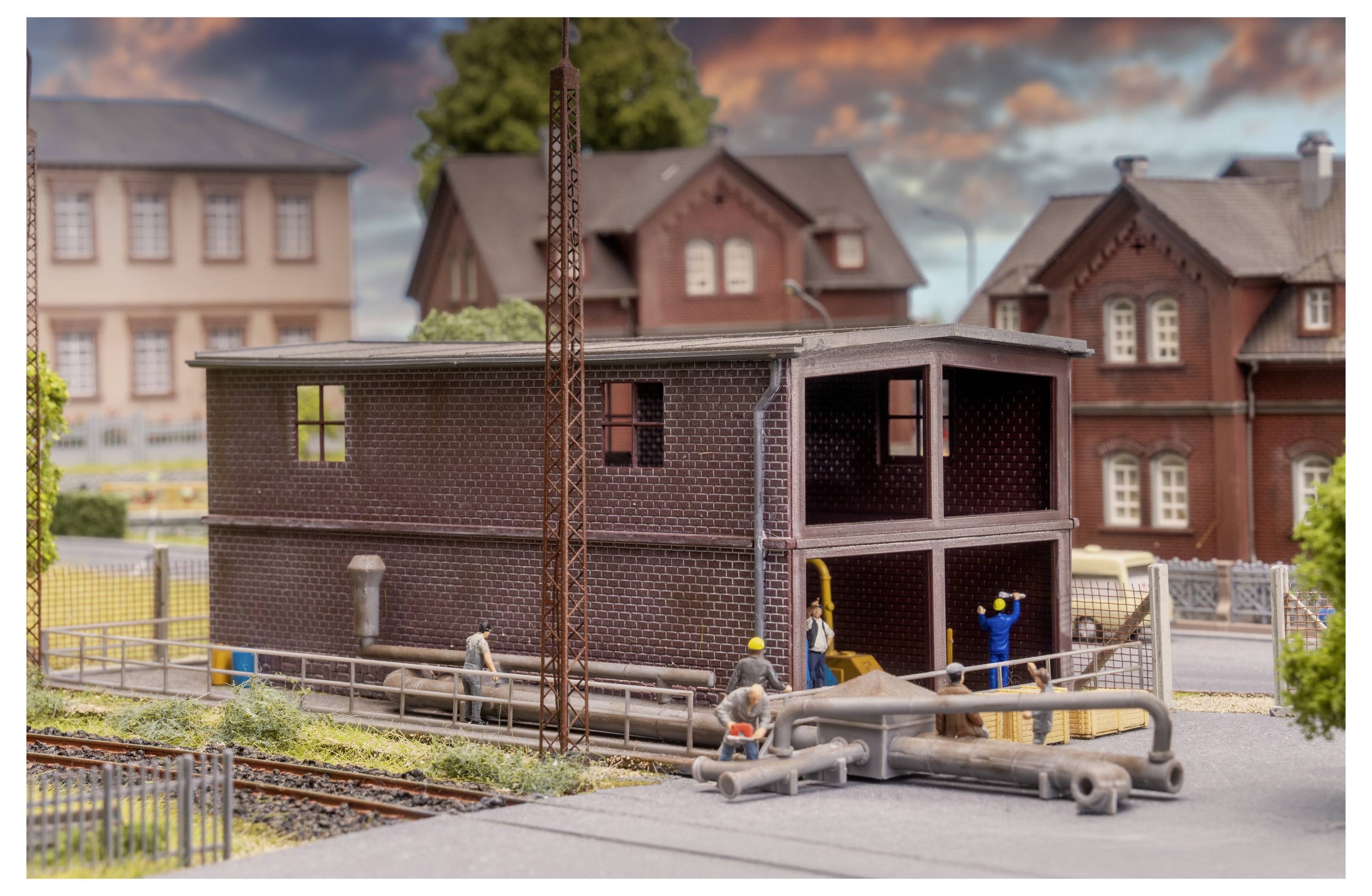 Modelleisenbahn-Industriegebäude mit Arbeitern und Rohrleitungen neben Eisenbahngleisen in einer Miniaturstadt-Szene.