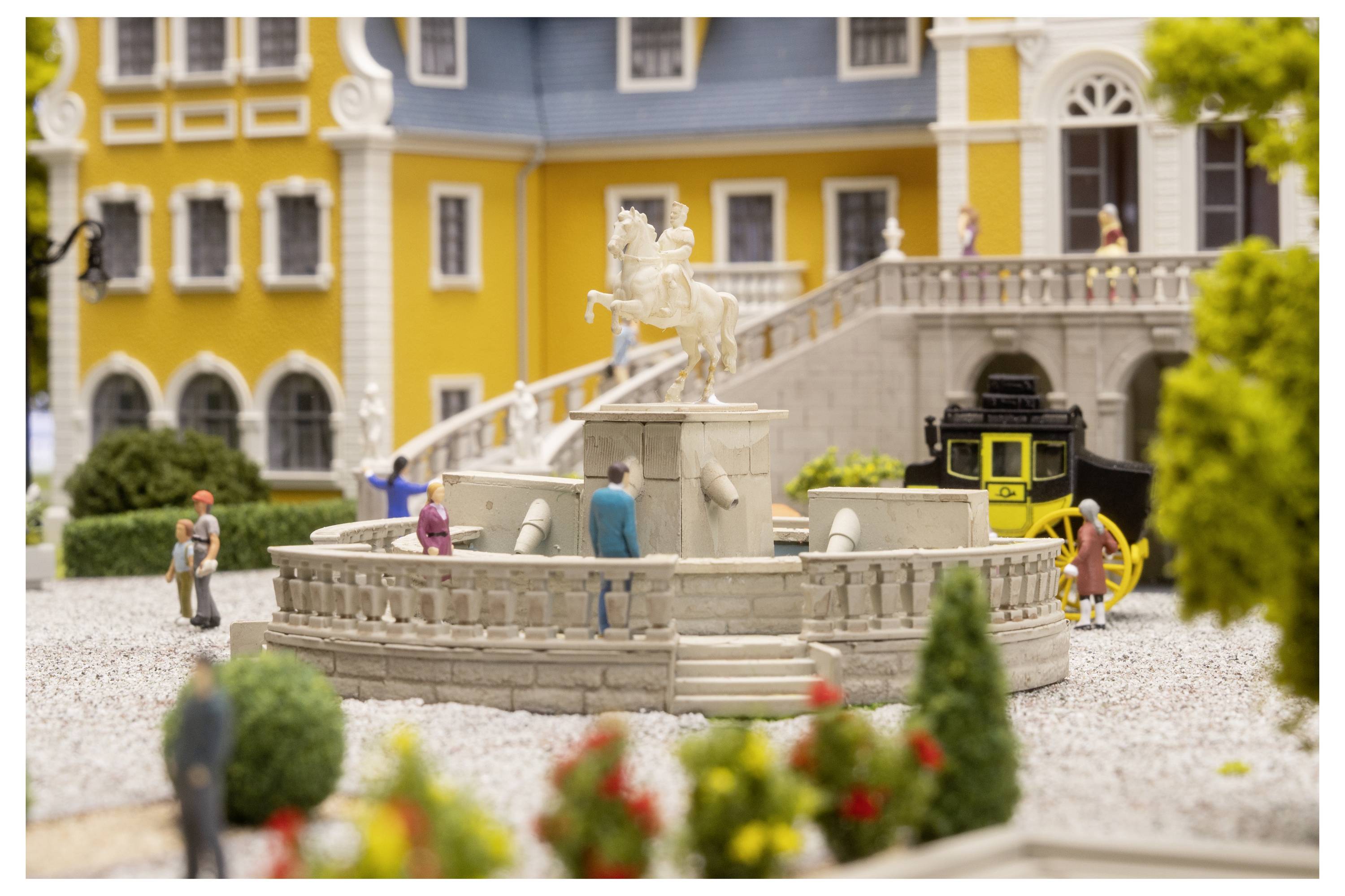 Miniatur-Außenszenerie mit einem zentralen Steinbrunnen und einer Statue, winzigen Menschen darum herum, und einer gelben Straßenbahn neben einem gelben Gebäude und einer grauen Treppe.