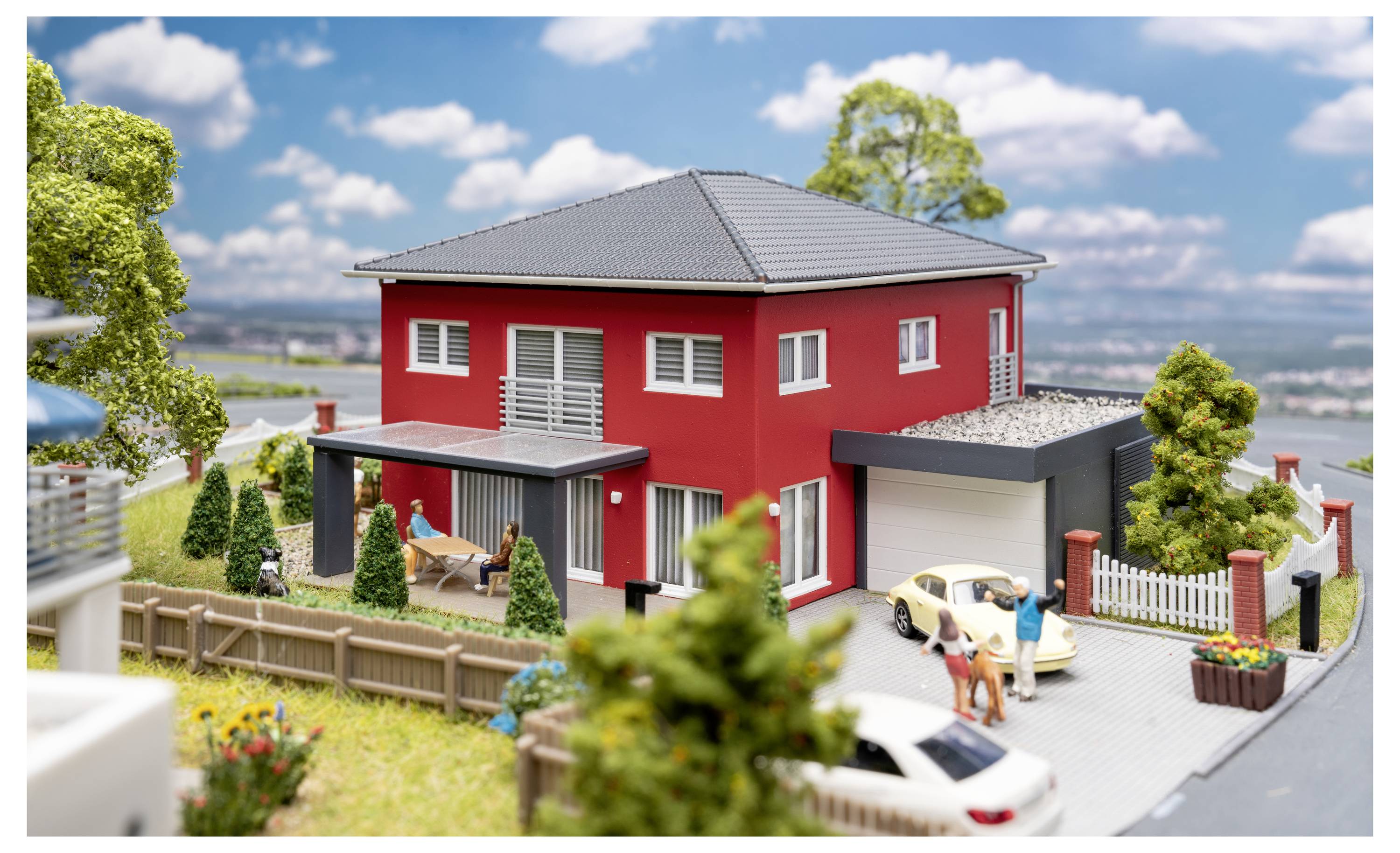 Modell eines modernen roten zweistöckigen Hauses mit eingezäuntem Hof, Terrasse, Einfahrt und kleinen Figuren neben geparkten Autos.