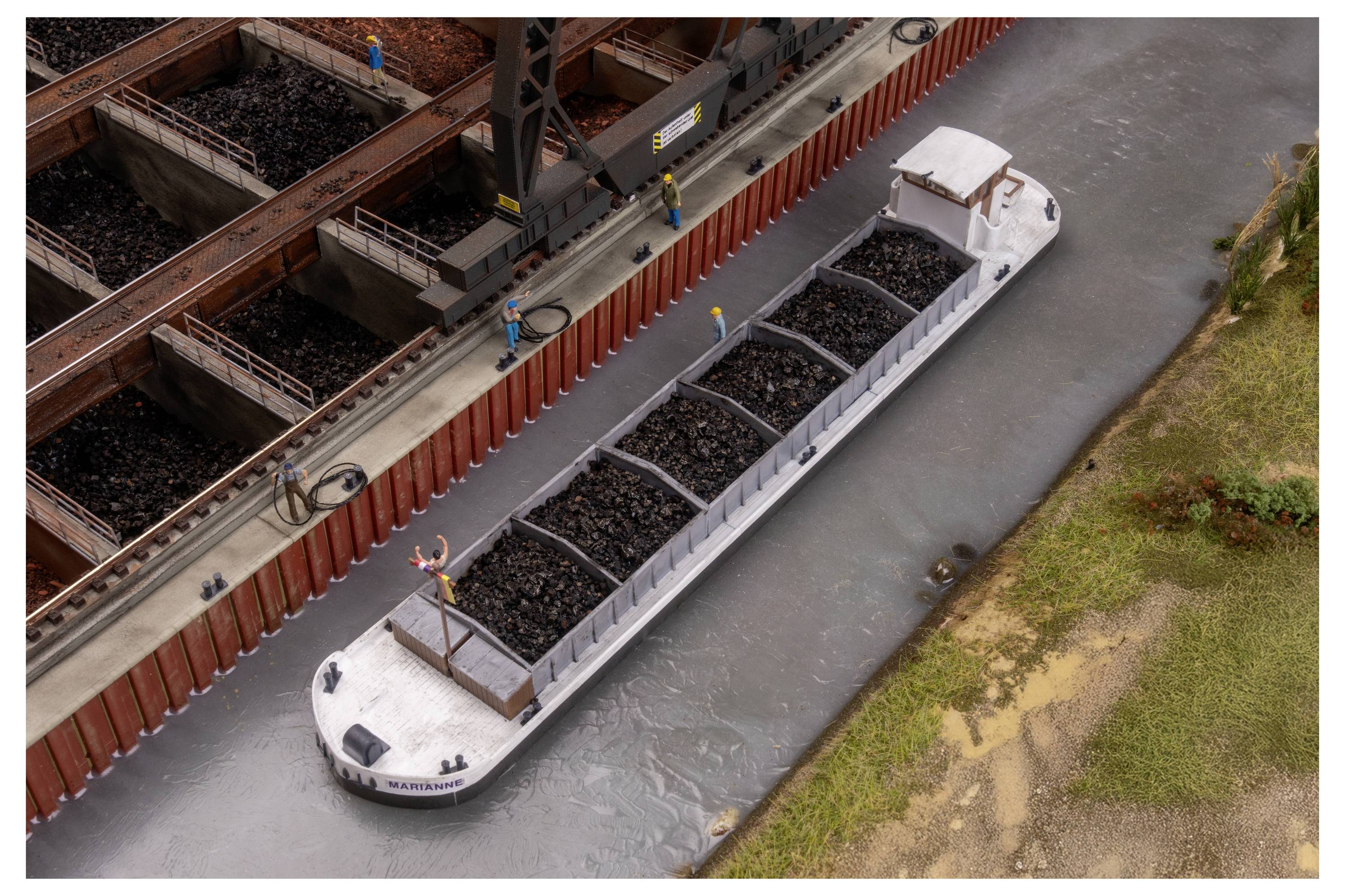 Eine mit Kohle beladene Industrieprahm bewegt sich durch einen Kanal neben einem Dock, mit Arbeitern auf dem Deck und Eisenbahnwagen, die in der Nähe entladen werden.