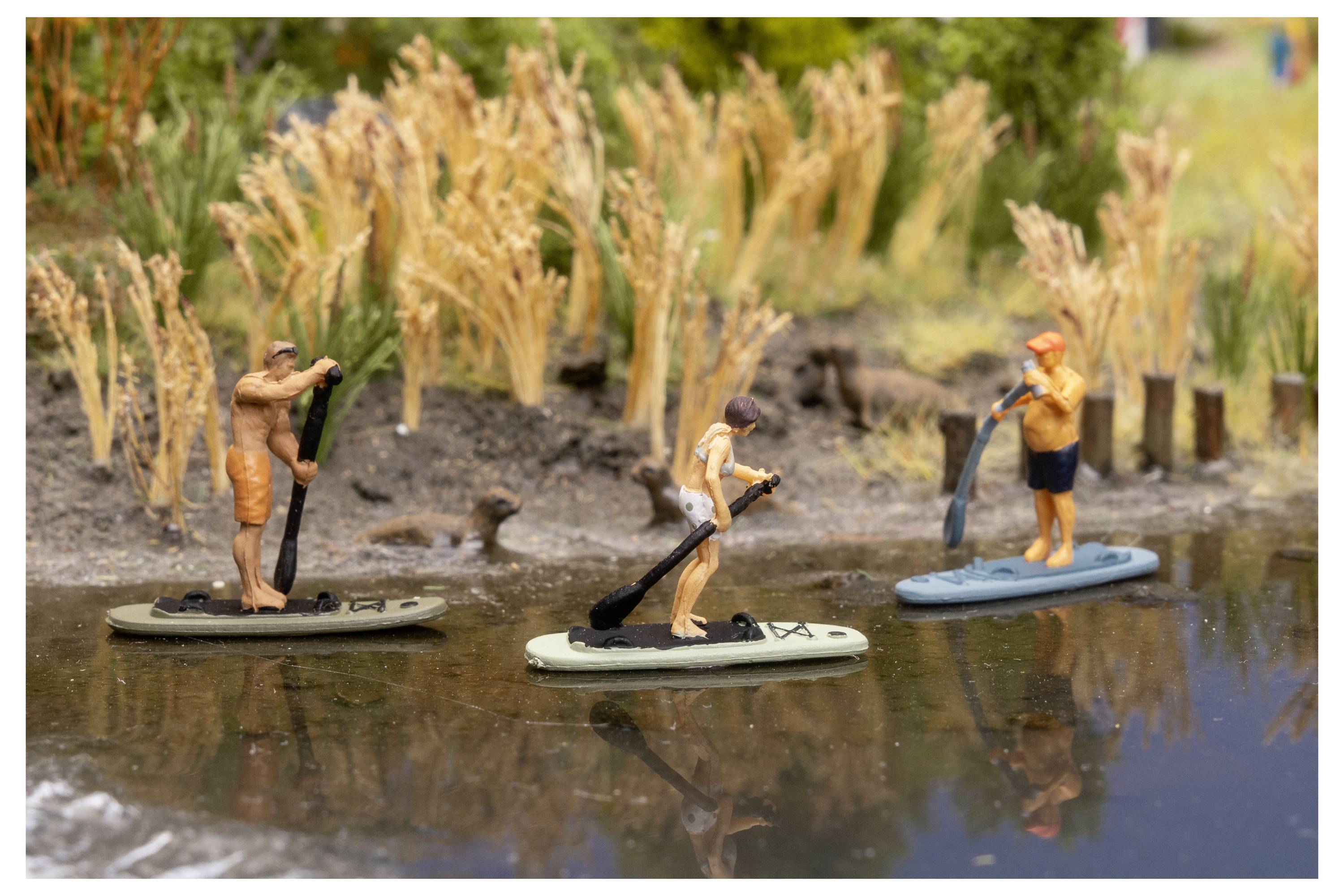 Drei winzige Personen stehen auf Paddleboards in flachem Wasser in der Nähe von Schilf und deuten auf eine Modellszene des Freizeitpaddelbrettsports hin.